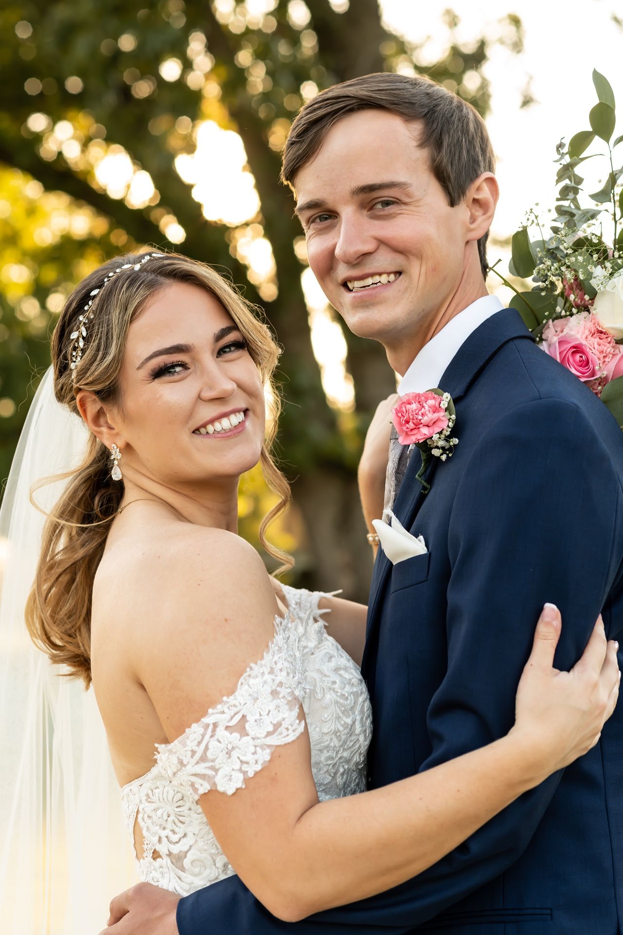 Newlywed couple embraces outdoors at sunset with bokeh lights and pink floral accents.