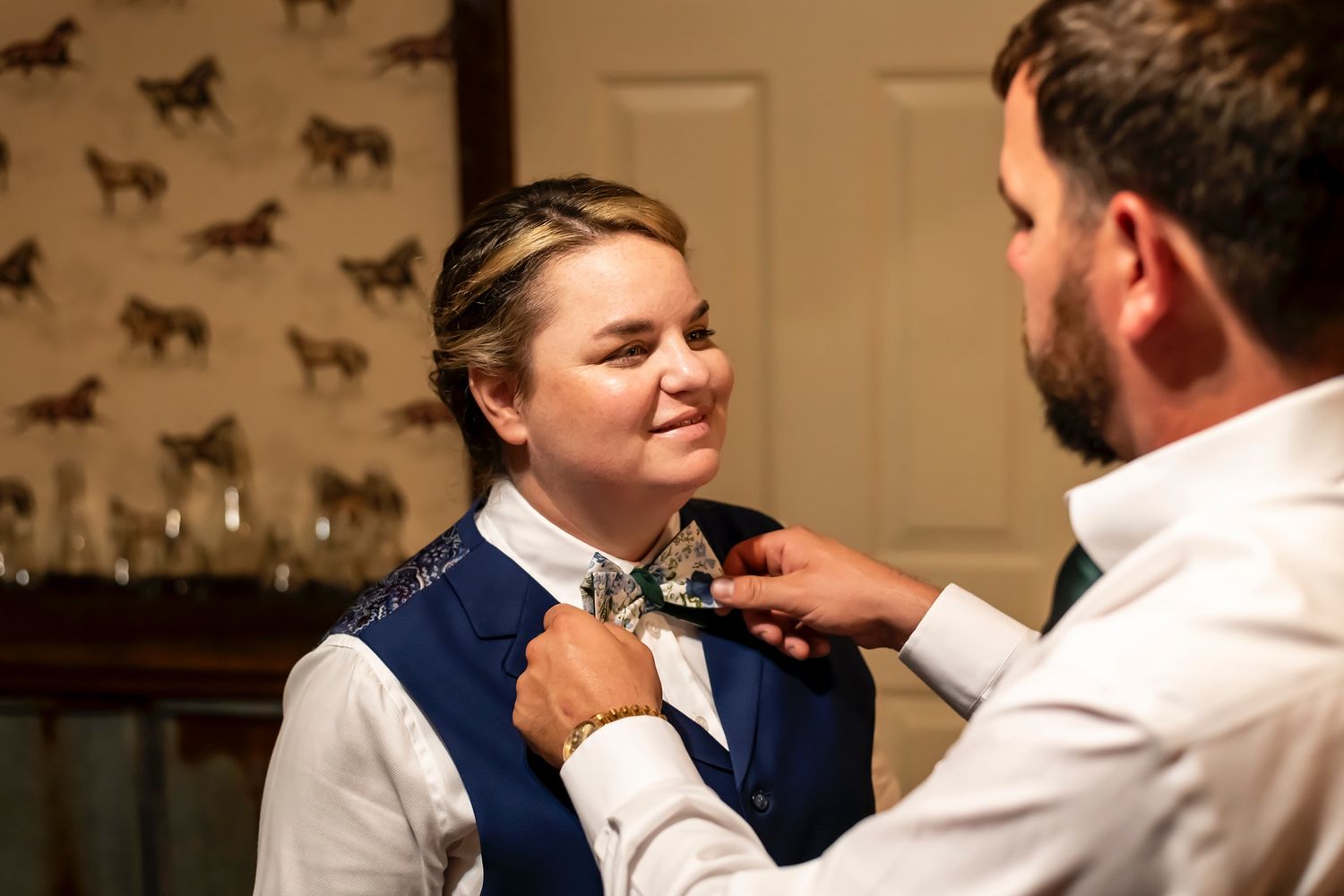 Person in a blue vest getting their tie adjusted by someone in a white shirt against a wall with horse silhouette pattern.