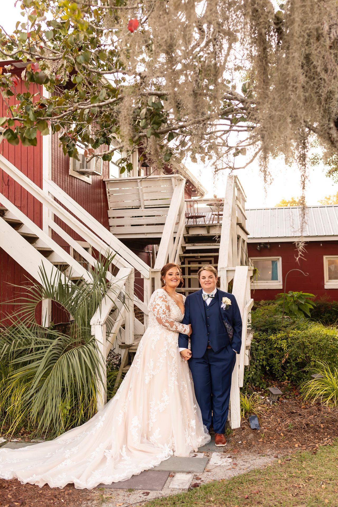 A couple stands together on white staircase outside red building with hanging Spanish moss at sunset.