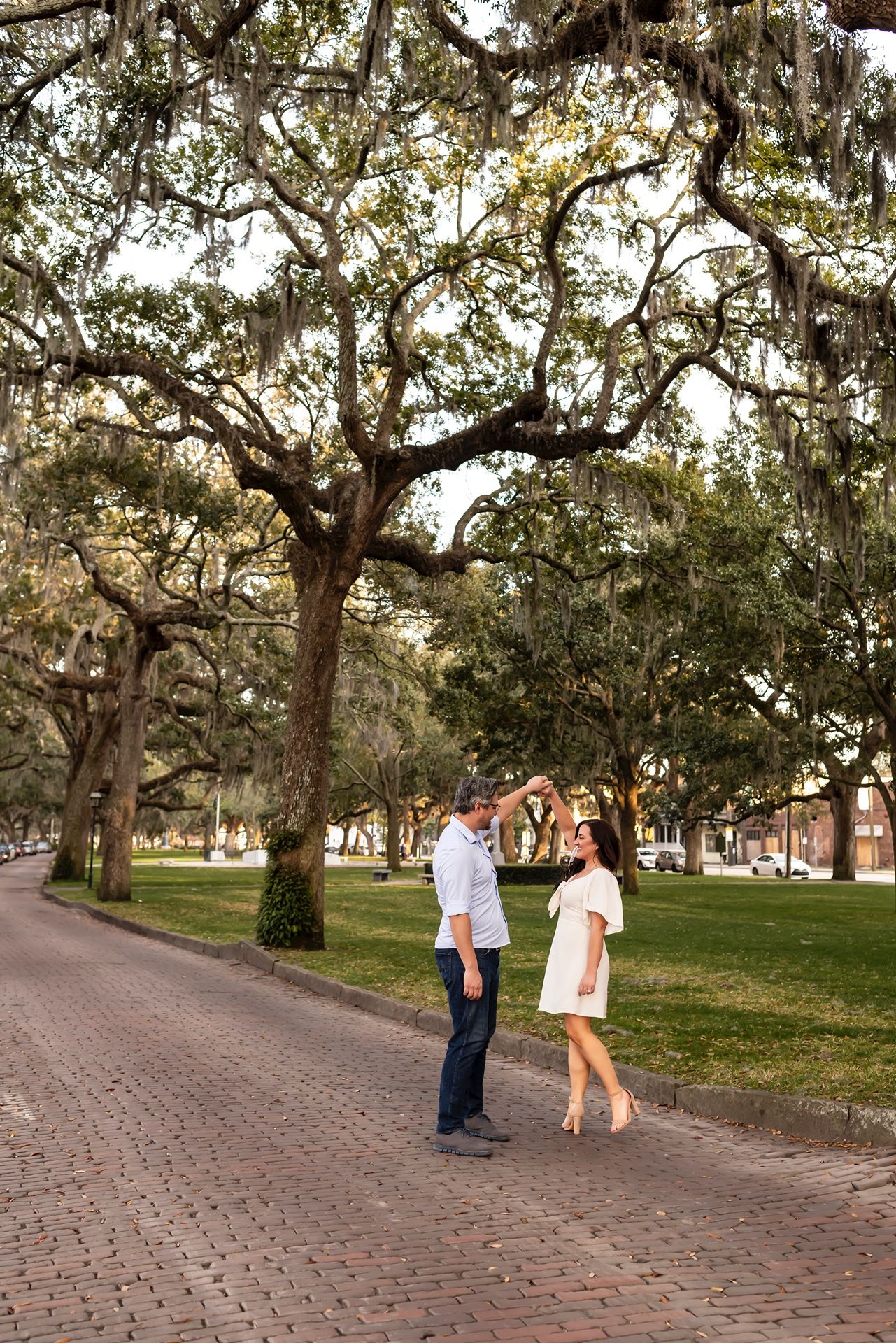 A romantic couple strolls together beneath sprawling oak trees draped with Spanish moss along a brick pathway.