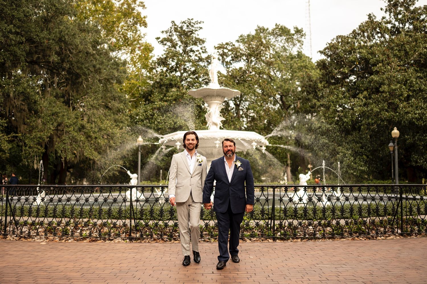 Wedding couple strolls down a scenic garden path with stone balustrades under green tree canopy on sunny day.