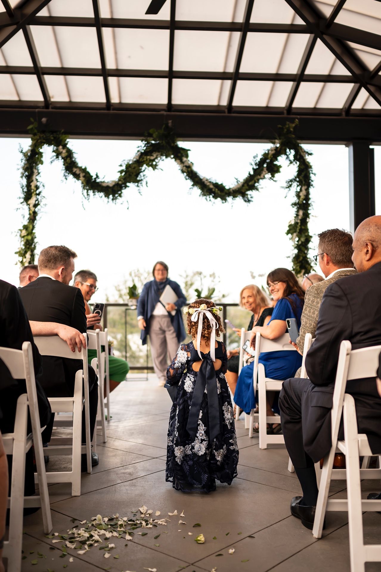 A small figure walks down the aisle during a wedding ceremony under a covered pavilion decorated with garlands.