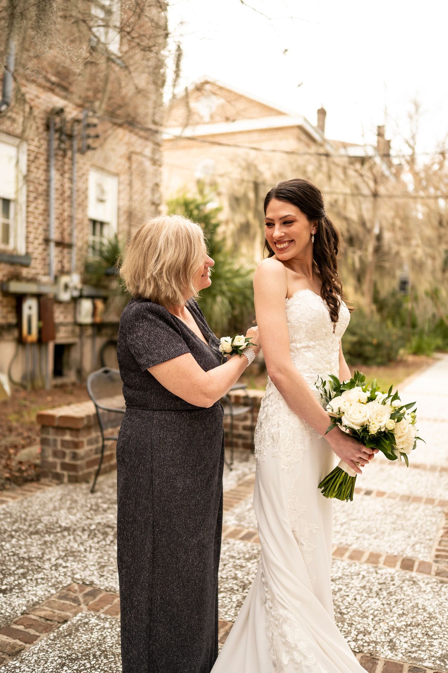 Person in elegant white lace wedding gown holding white bouquet while being helped with dress in outdoor courtyard setting.