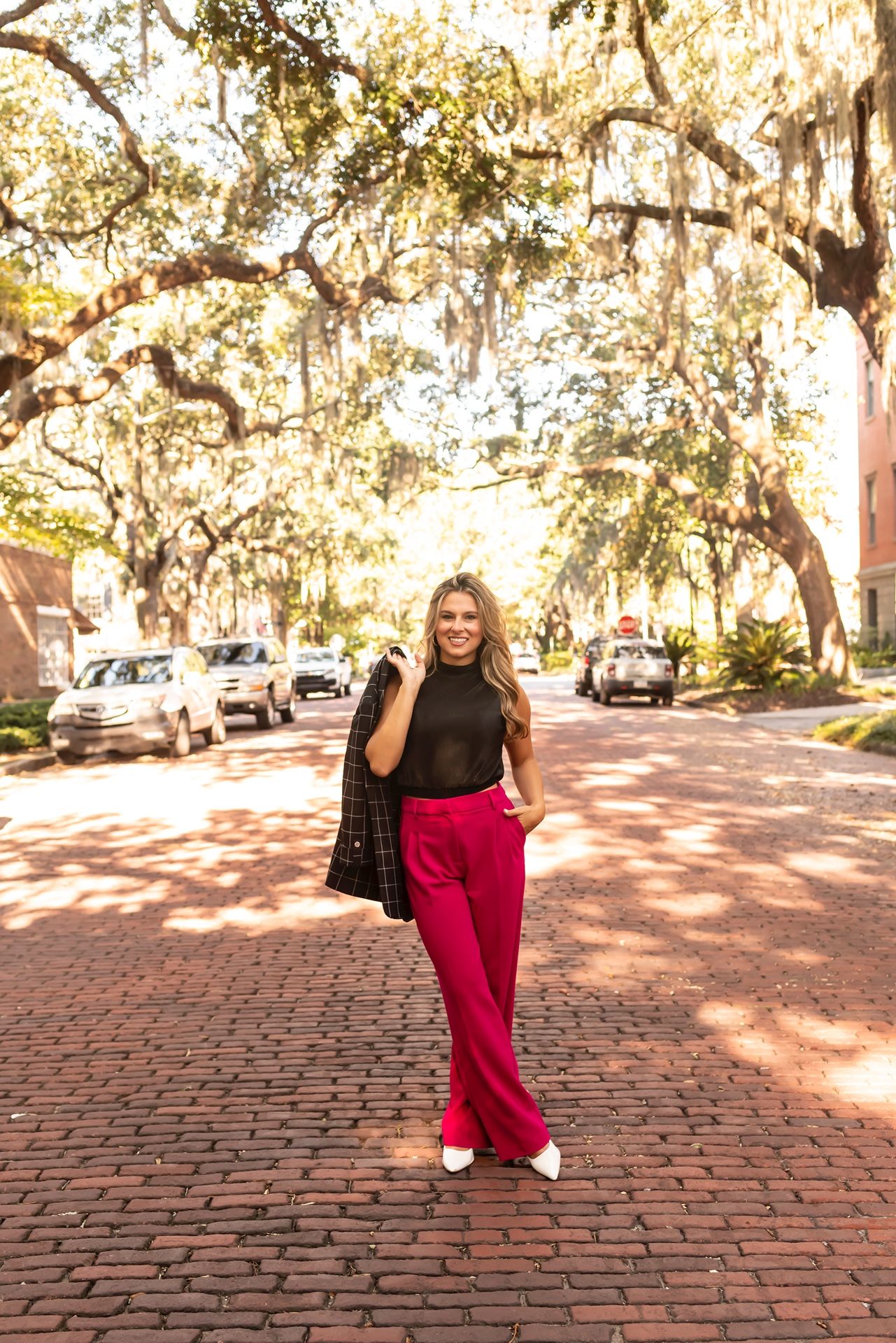 Red pants and black top outfit shown in a series of poses on a sunlit brick courtyard with moss-covered oak trees.