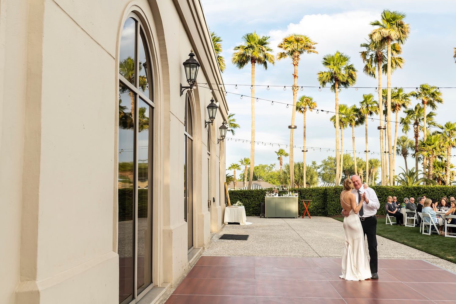 A couple shares a romantic moment on a terrace surrounded by palm trees and elegant white columns at sunset.