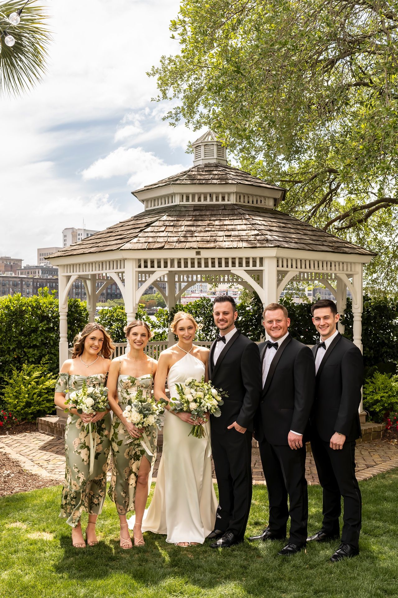 Wedding party poses on a garden lawn in front of a white gazebo during an elegant outdoor ceremony.