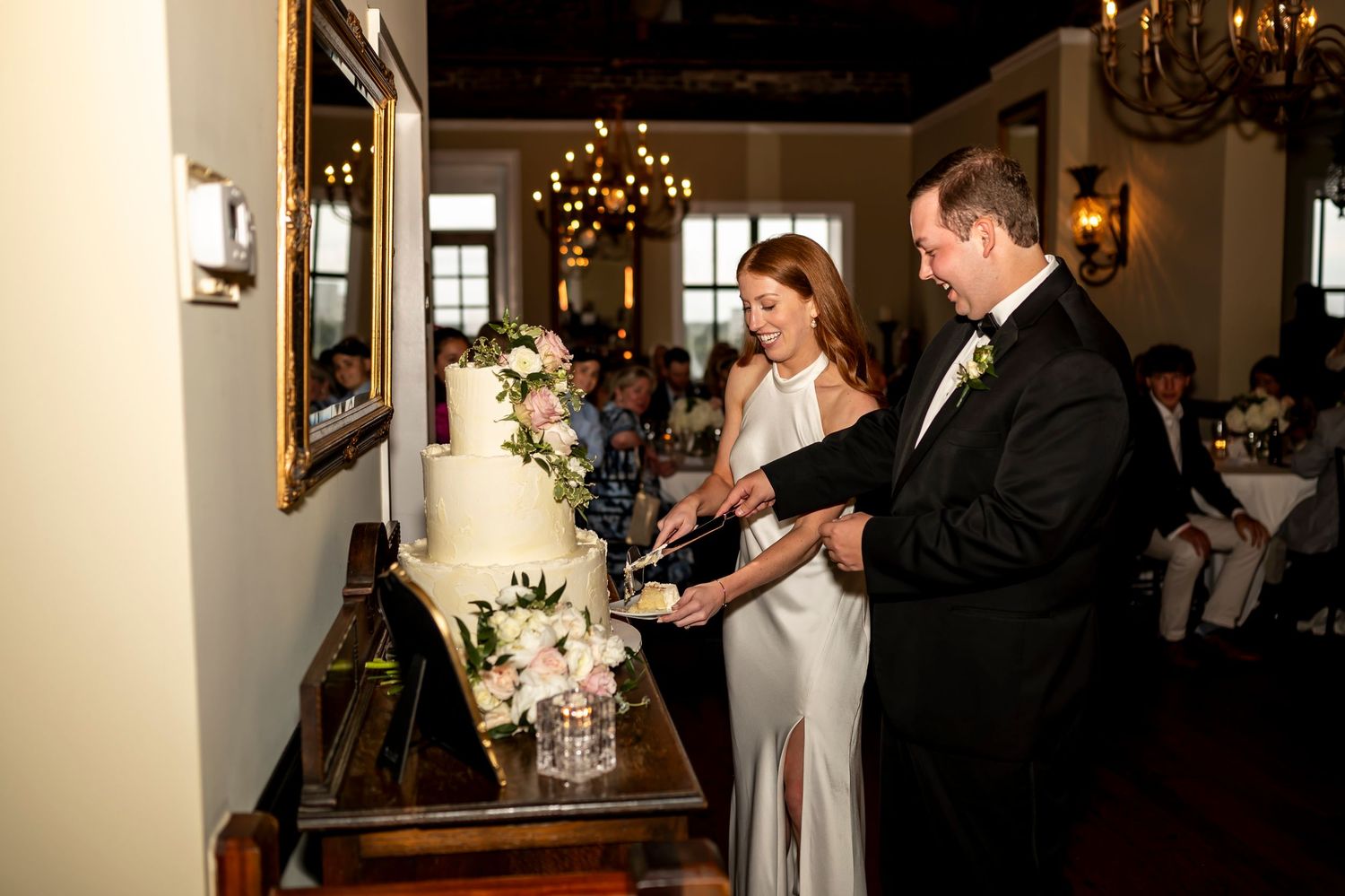 A couple cuts their elegant white floral wedding cake together in a dimly lit reception venue with chandeliers.
