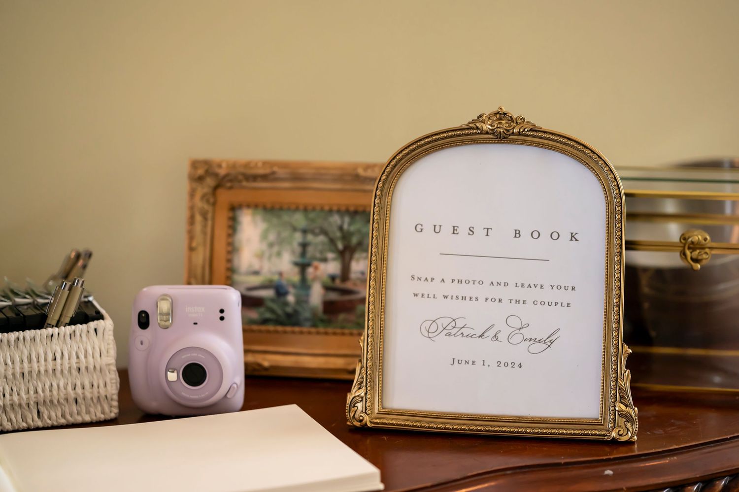 A vintage-style gold frame displays a guest book sign next to a pink Instax camera on a table.