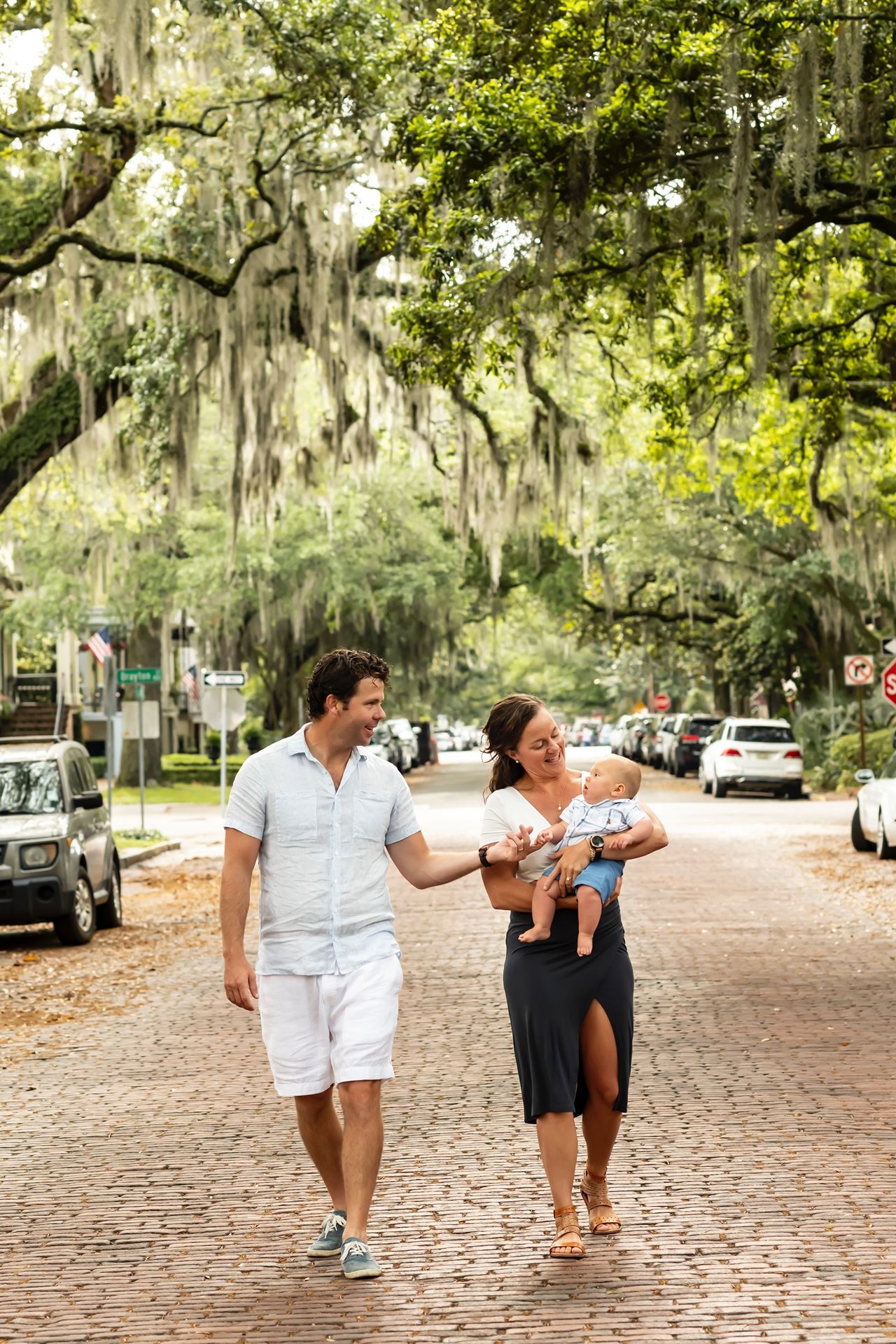 A couple walks with a baby on a scenic brick street lined with Spanish moss-draped oak trees in the South.