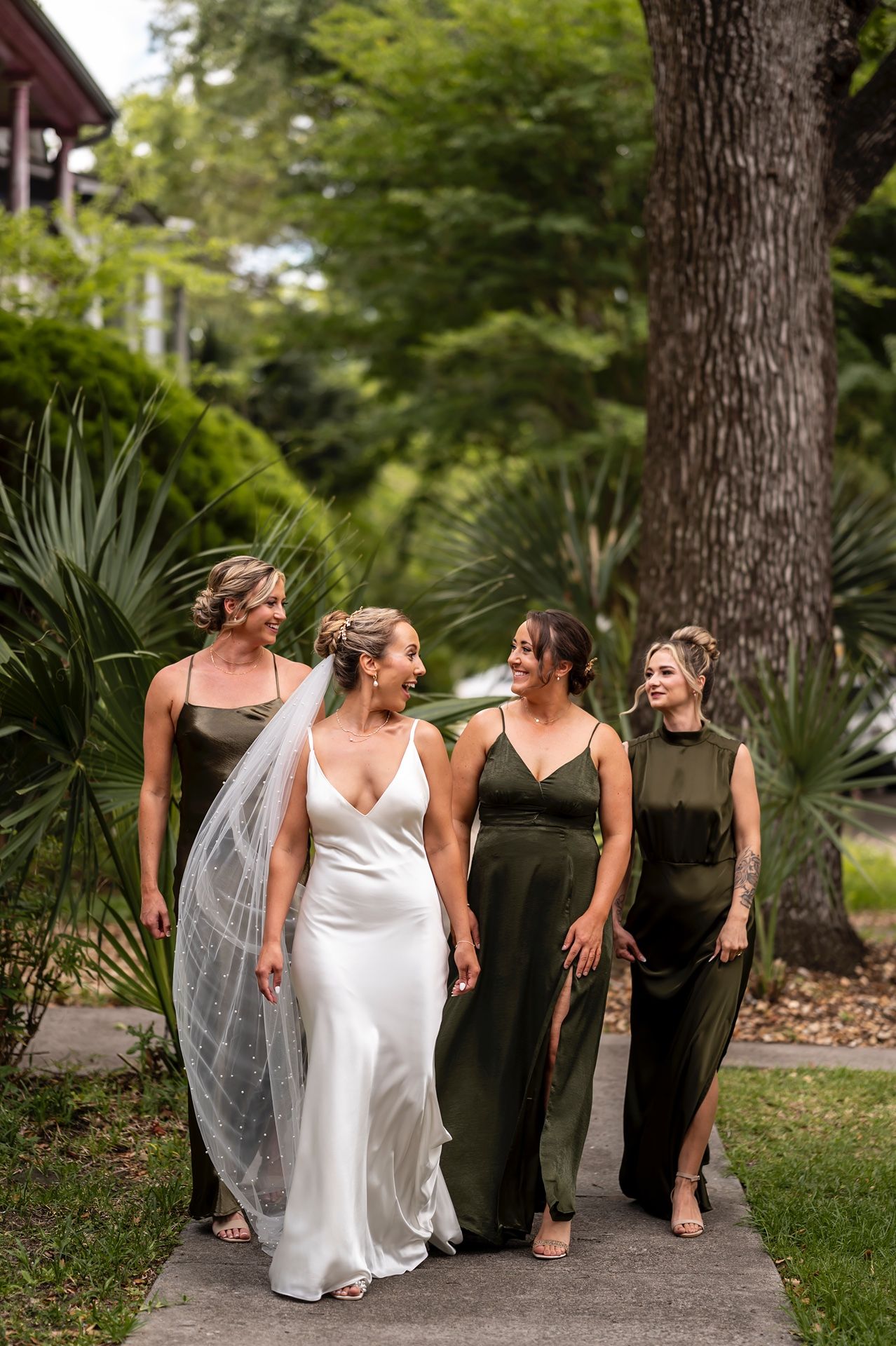 Wedding party walks together on garden path surrounded by lush greenery and palm trees in outdoor setting.