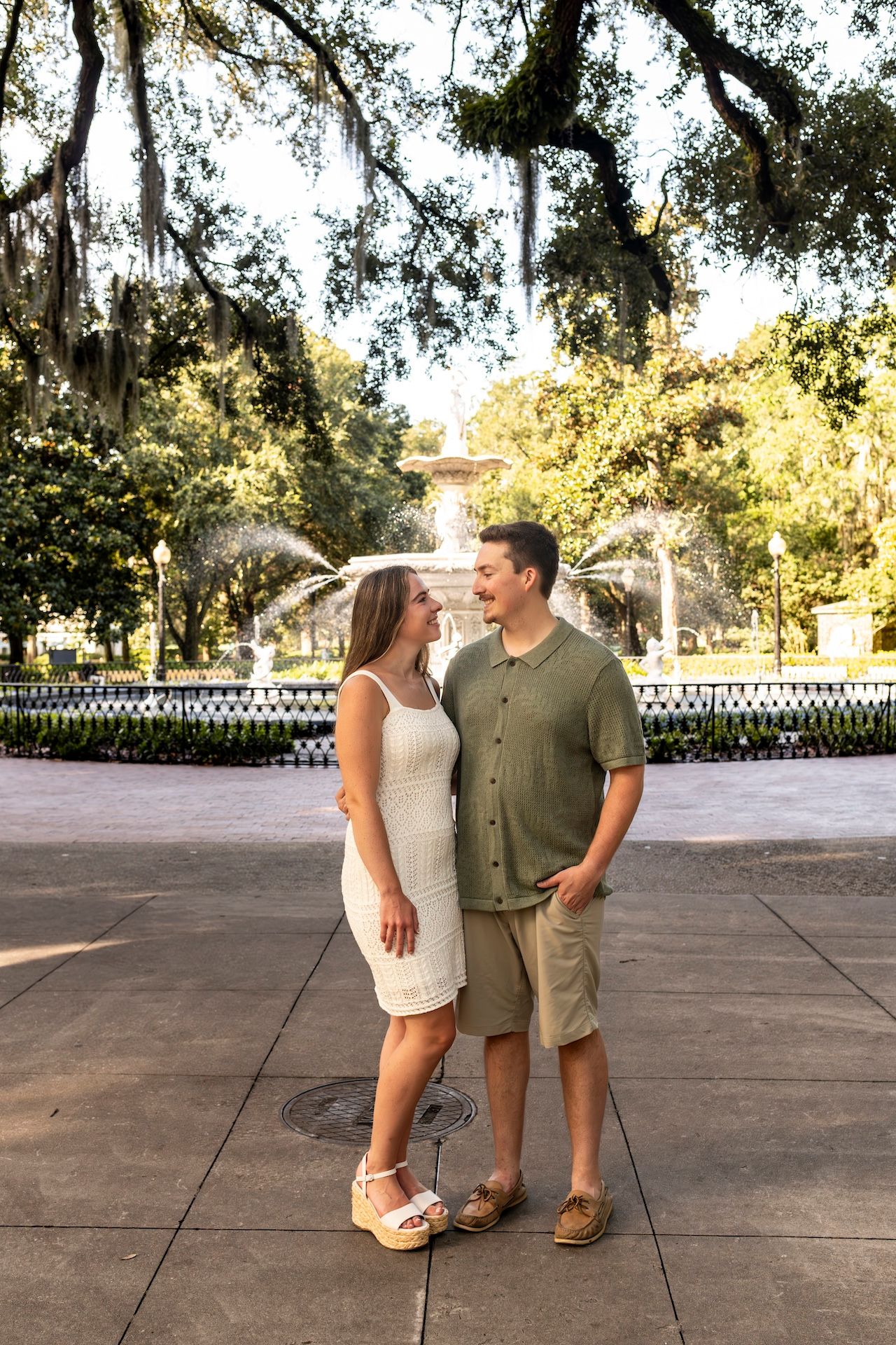 A couple shares a romantic moment on a paved outdoor pathway beneath mossy oak trees and soft sunlight.