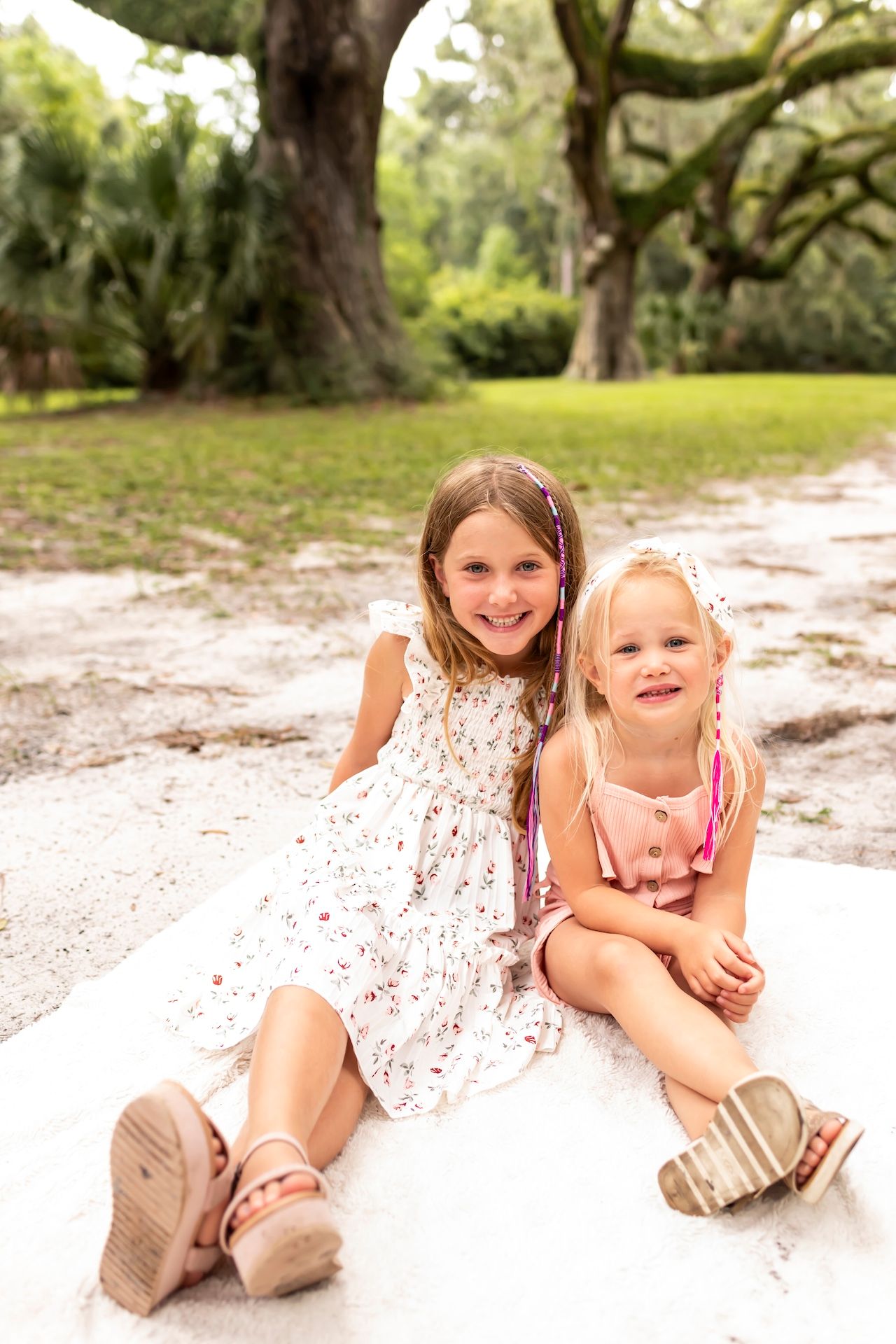 Two young ones lay playfully on white sand beneath majestic oak trees in a park setting.
