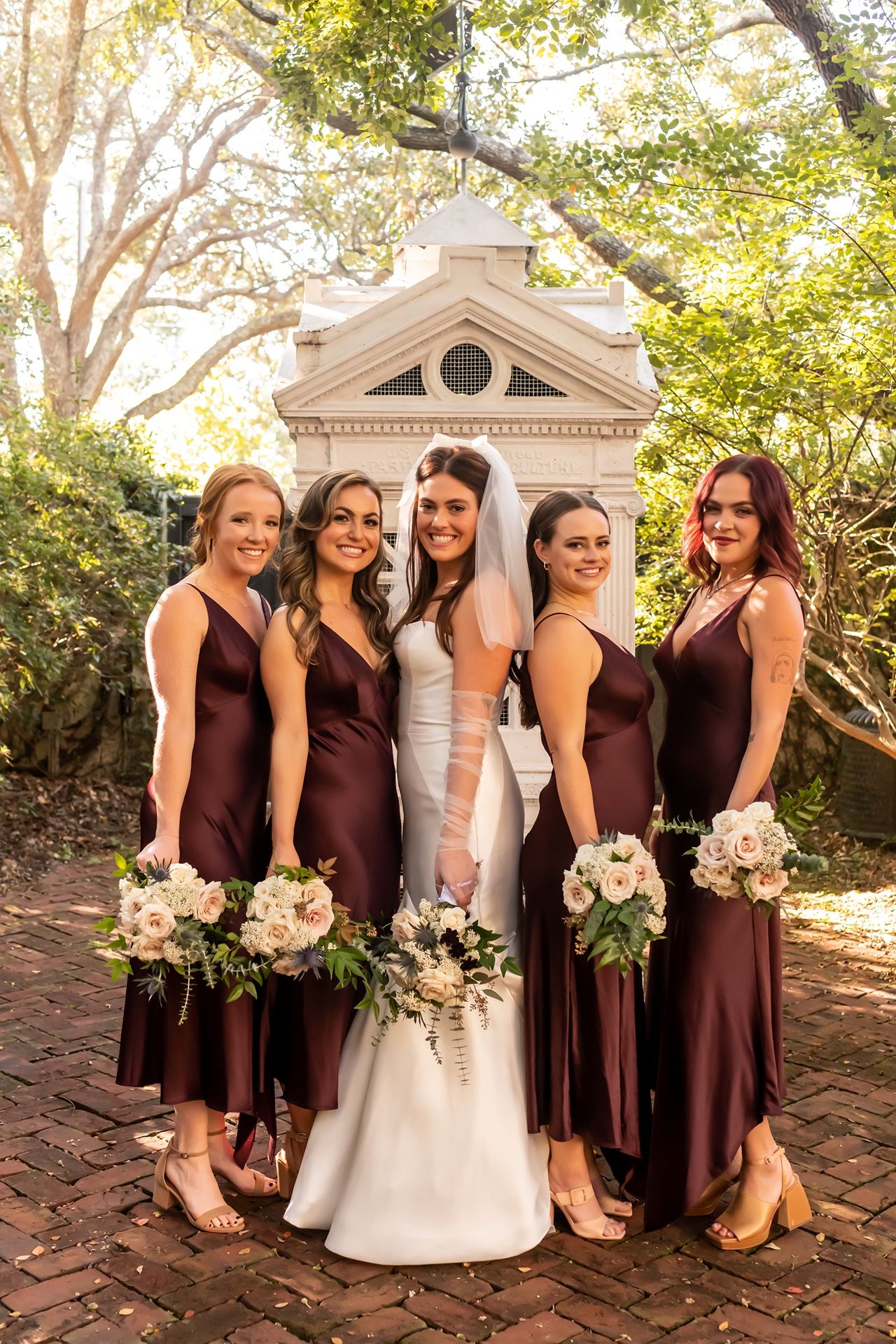 Wedding party in burgundy bridesmaid dresses and white bridal gown posing together in front of small chapel gazebo outdoors.
