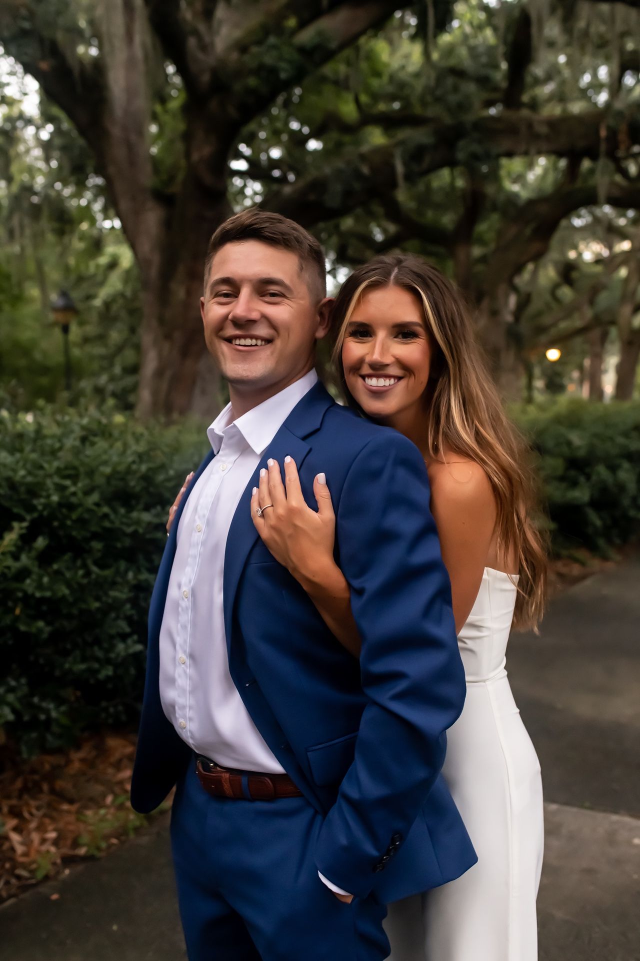 A couple in wedding attire pose on a tree-lined path, with the bride in white and groom in navy blue suit.