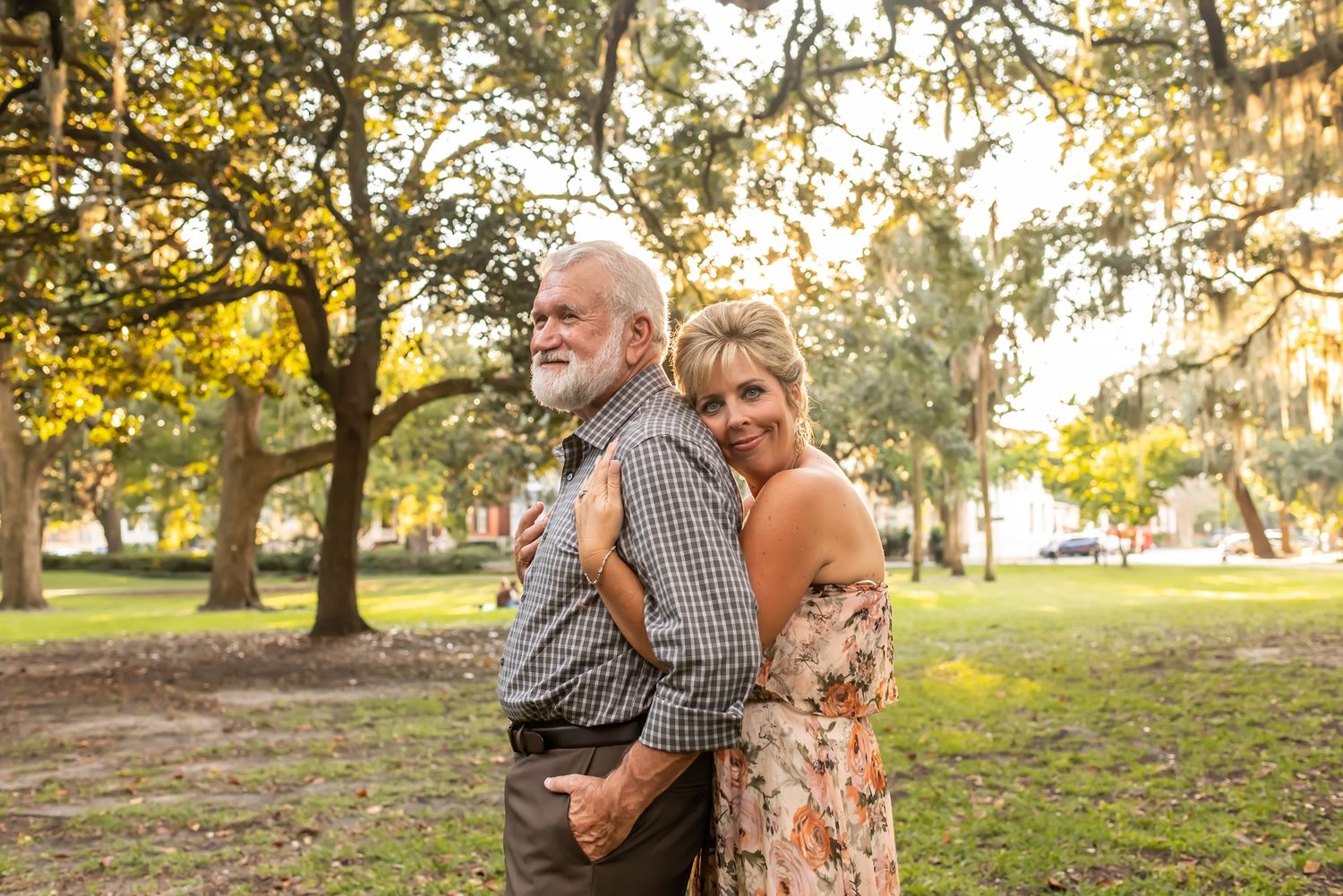 Two people embrace while standing in a sunlit park with moss-draped oak trees in the background.