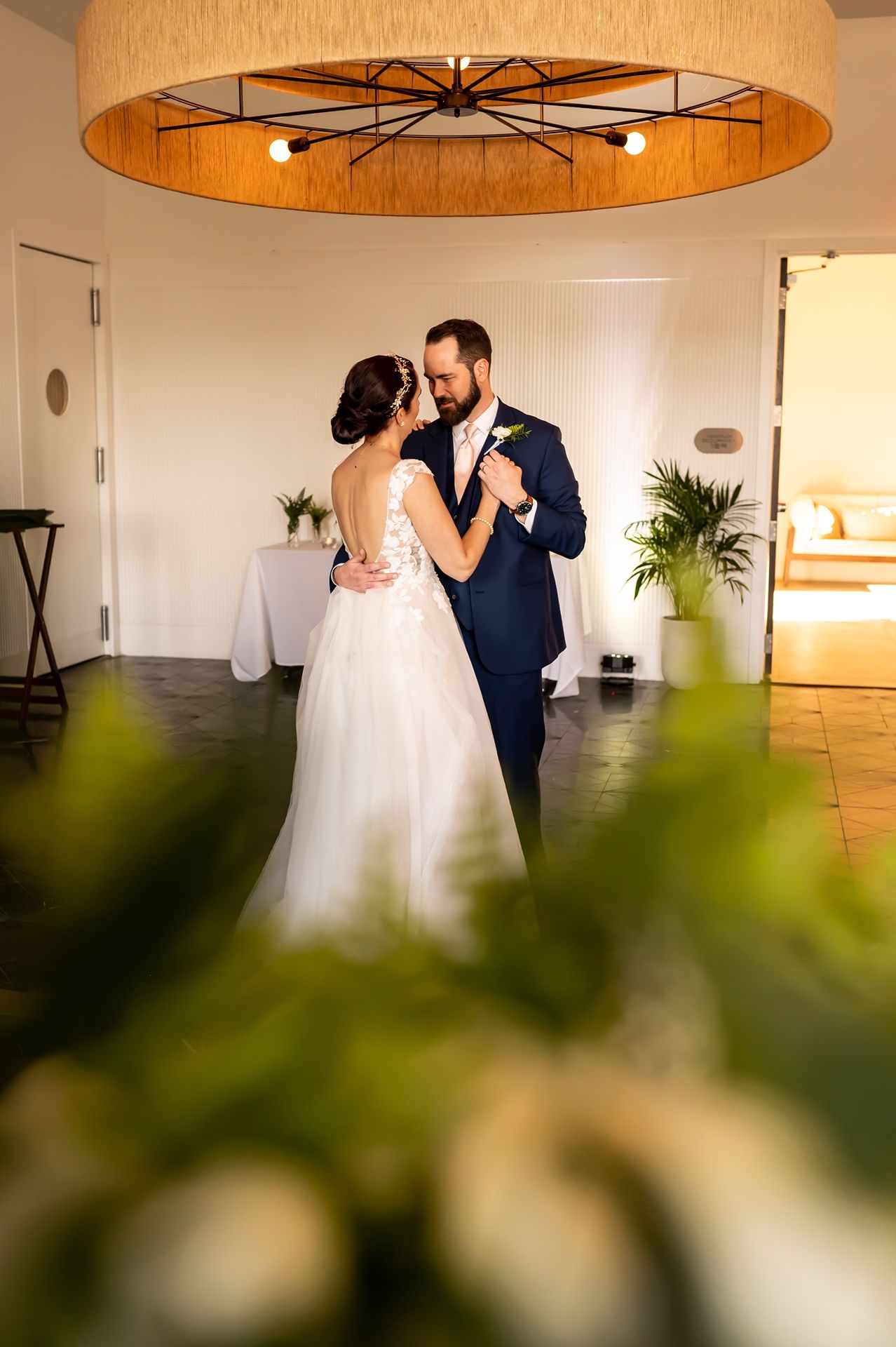 Wedding couple shares intimate dance moment under wooden circular chandelier in elegant reception venue.