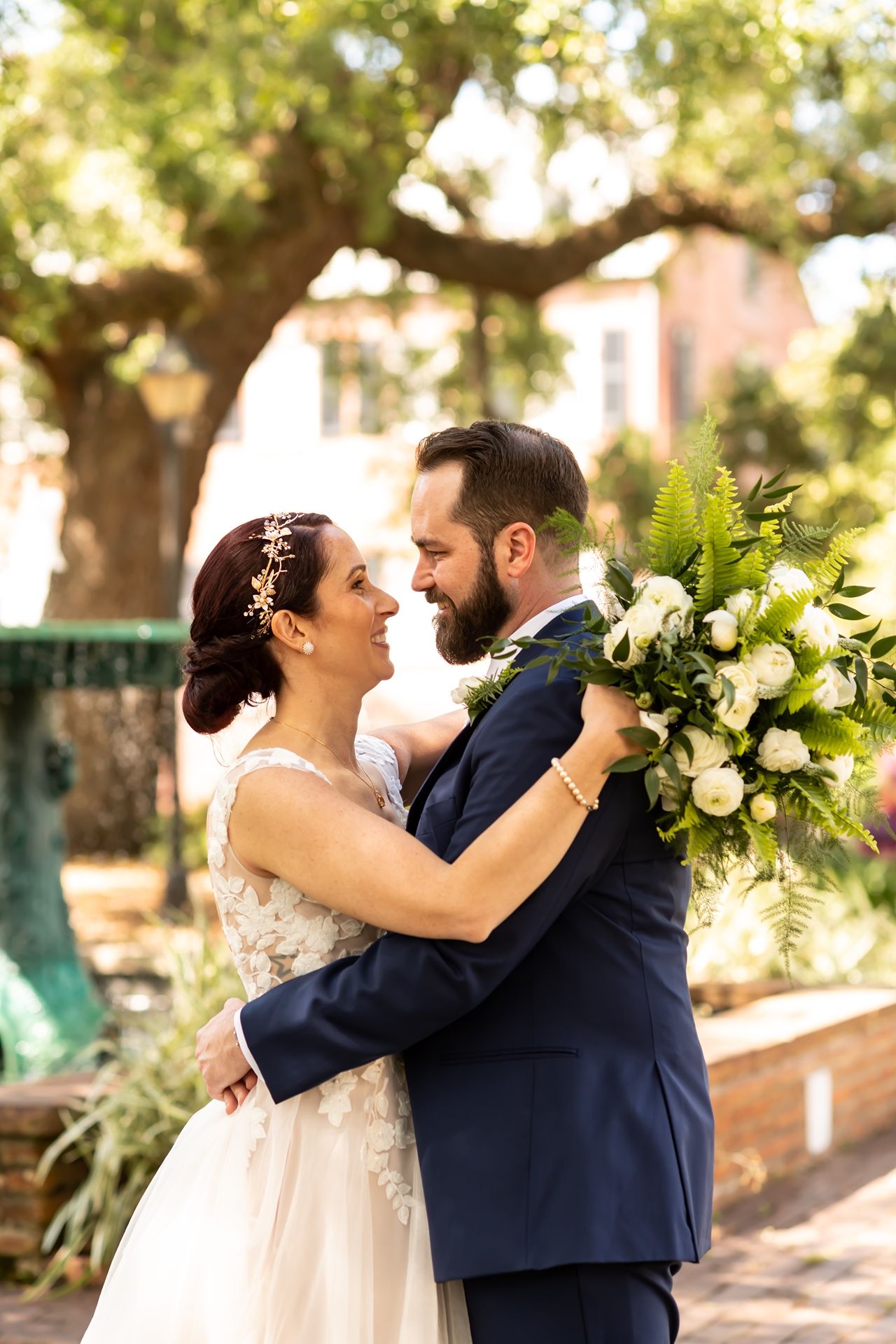 A joyful wedding moment captured outdoors with a beautiful white bouquet and navy suit under a sunlit tree canopy.