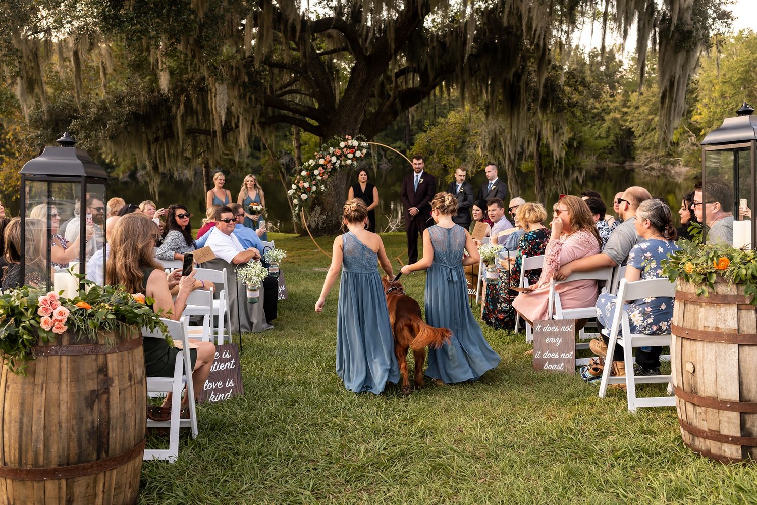A rustic outdoor wedding ceremony takes place under moss-draped oak trees with white chairs arranged on the lawn.