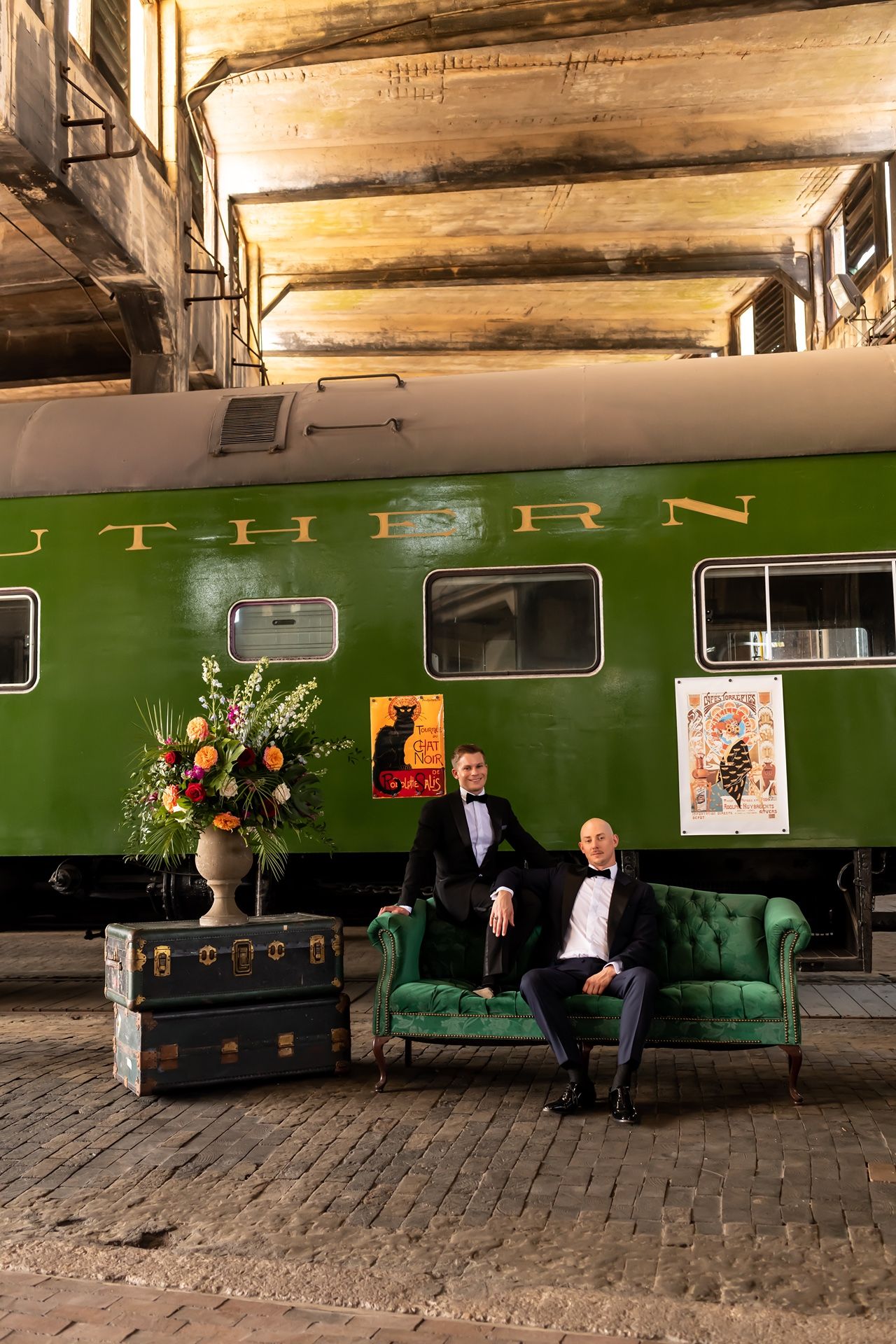 Two men sitting on a vintage green couch next to a retro train car with decorative flowers and posters.