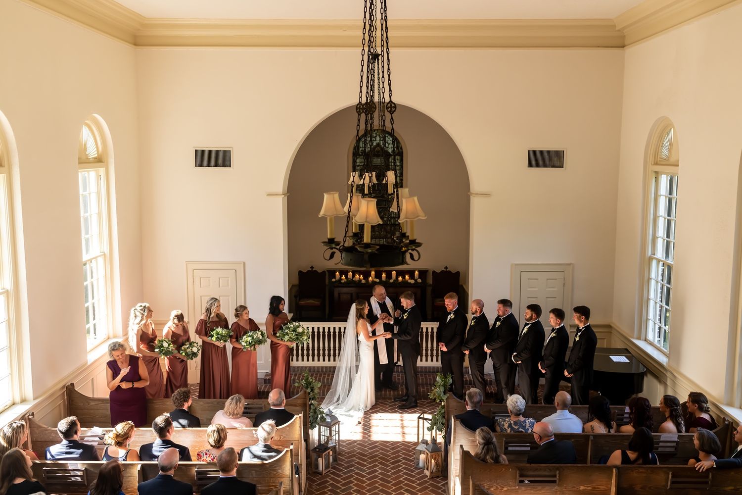 Wedding ceremony in a traditional church chapel with bridal party and groomsmen standing at the altar under chandelier.
