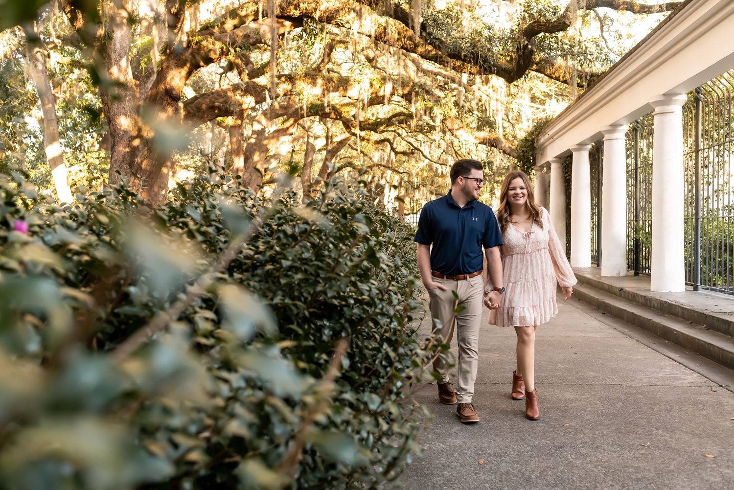 A couple strolls hand in hand along a covered walkway with sunlit foliage overhead creating a romantic atmosphere.