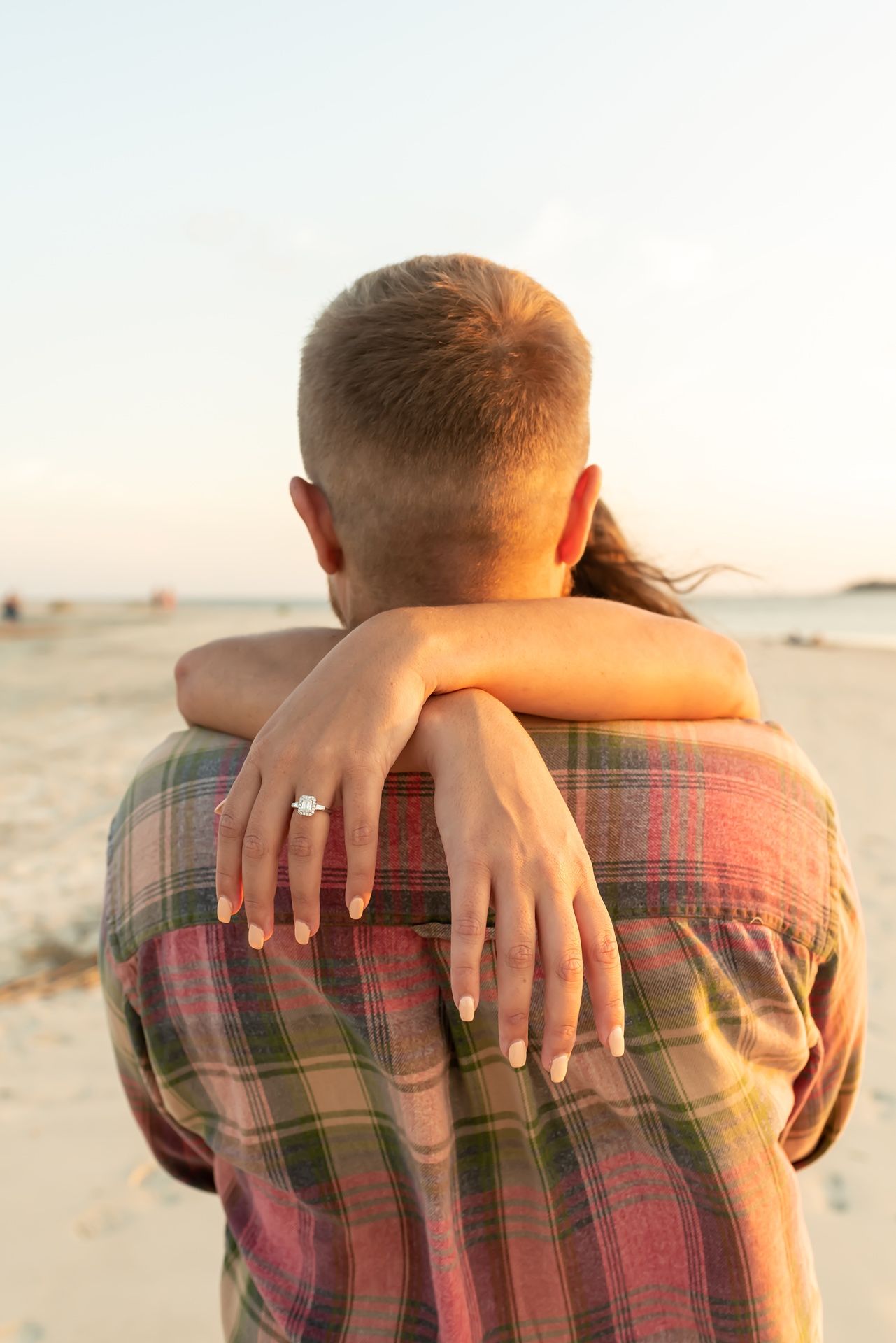 A couple embraces on a beach at sunset, with a sparkling engagement ring visible on the woman's hand.