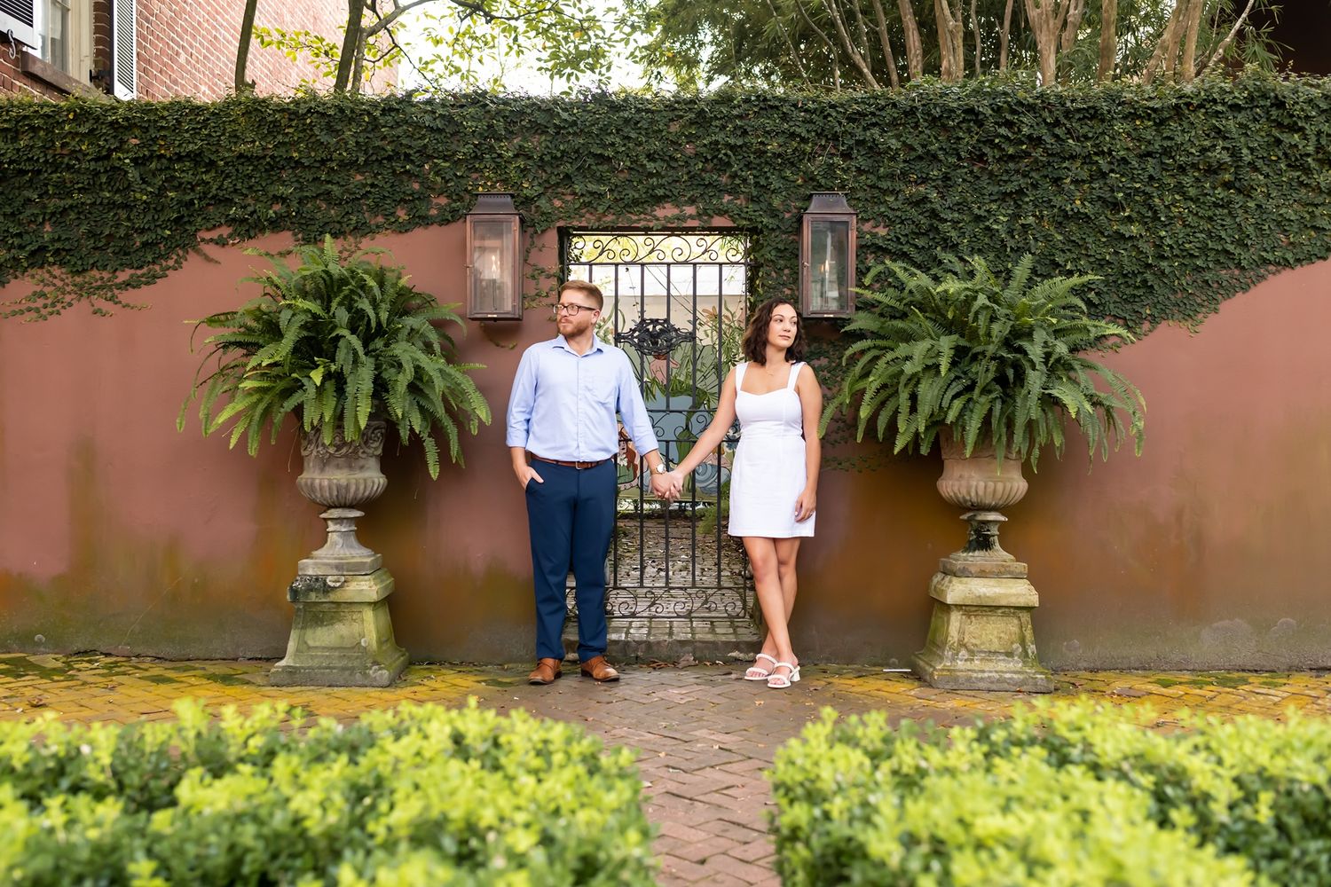 A couple poses in front of a rustic terracotta wall with ivy and potted ferns at a garden entrance.
