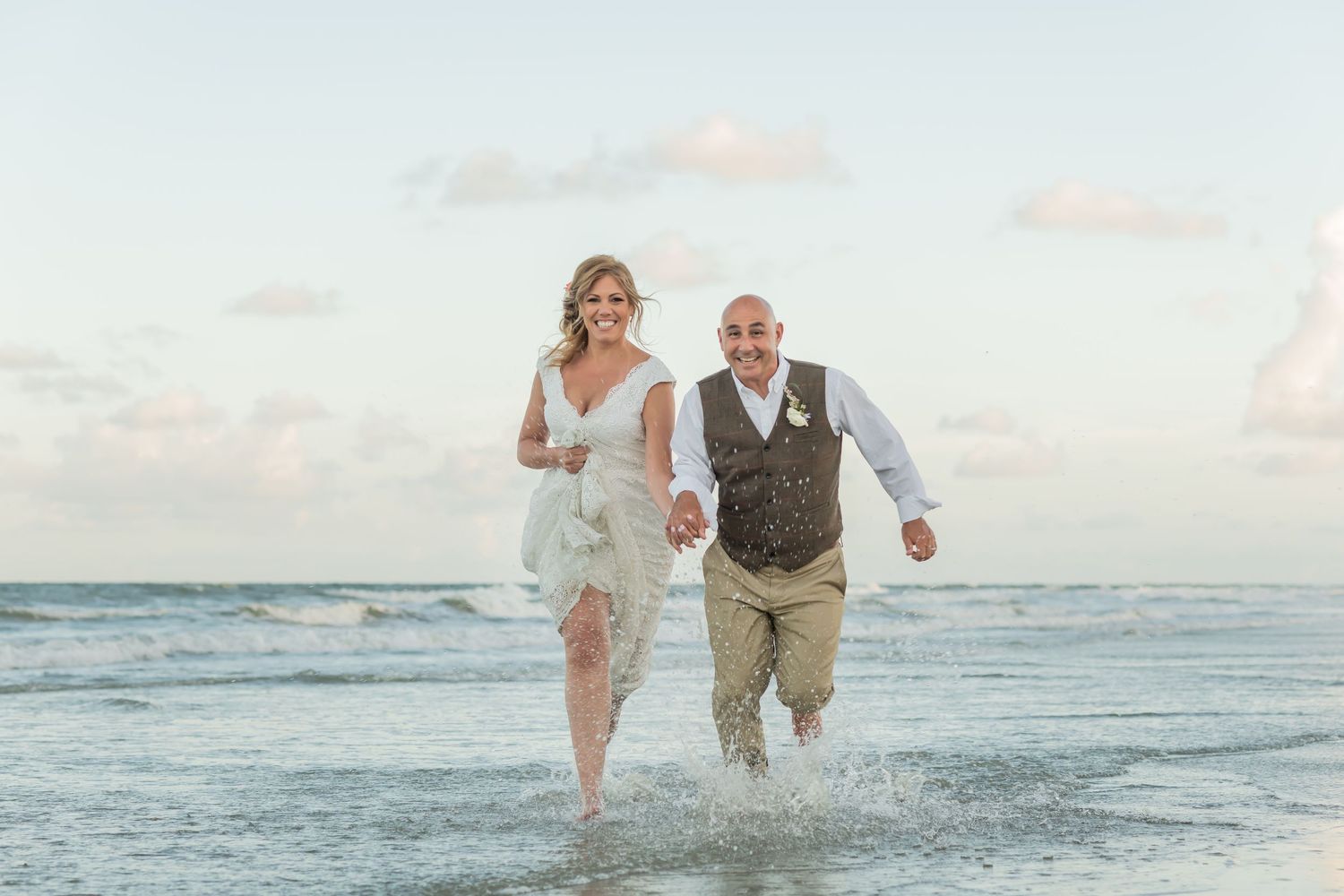A couple in wedding attire runs joyfully through shallow ocean waves at sunset on a beach.