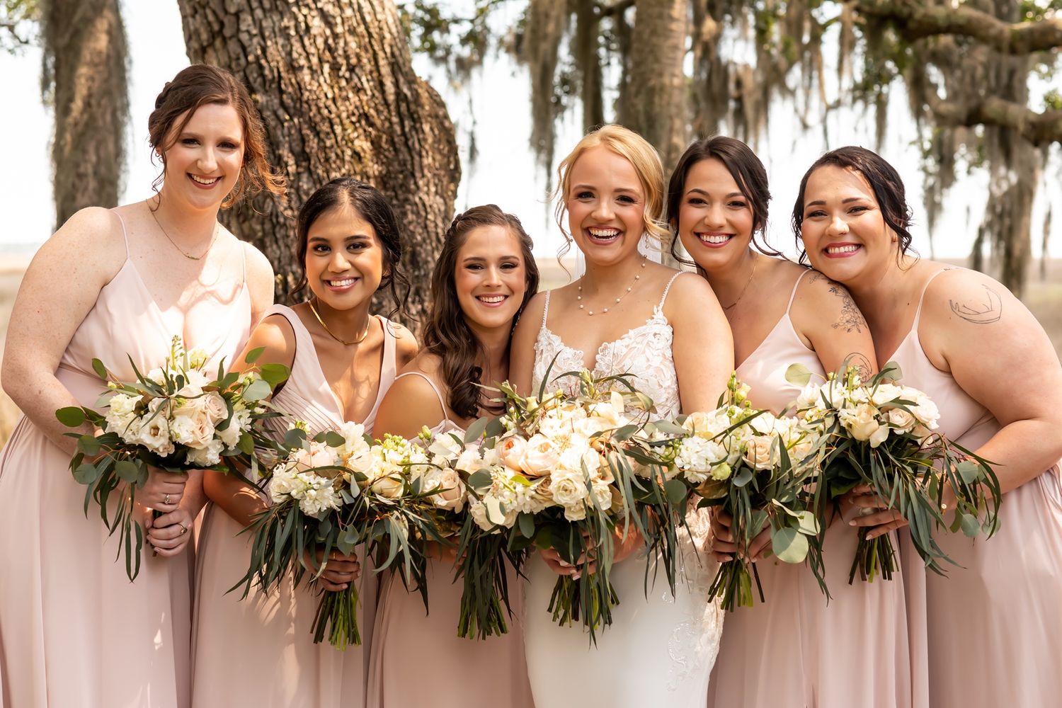 Bridesmaids in blush pink dresses hold lush white and green floral bouquets while standing together outdoors.