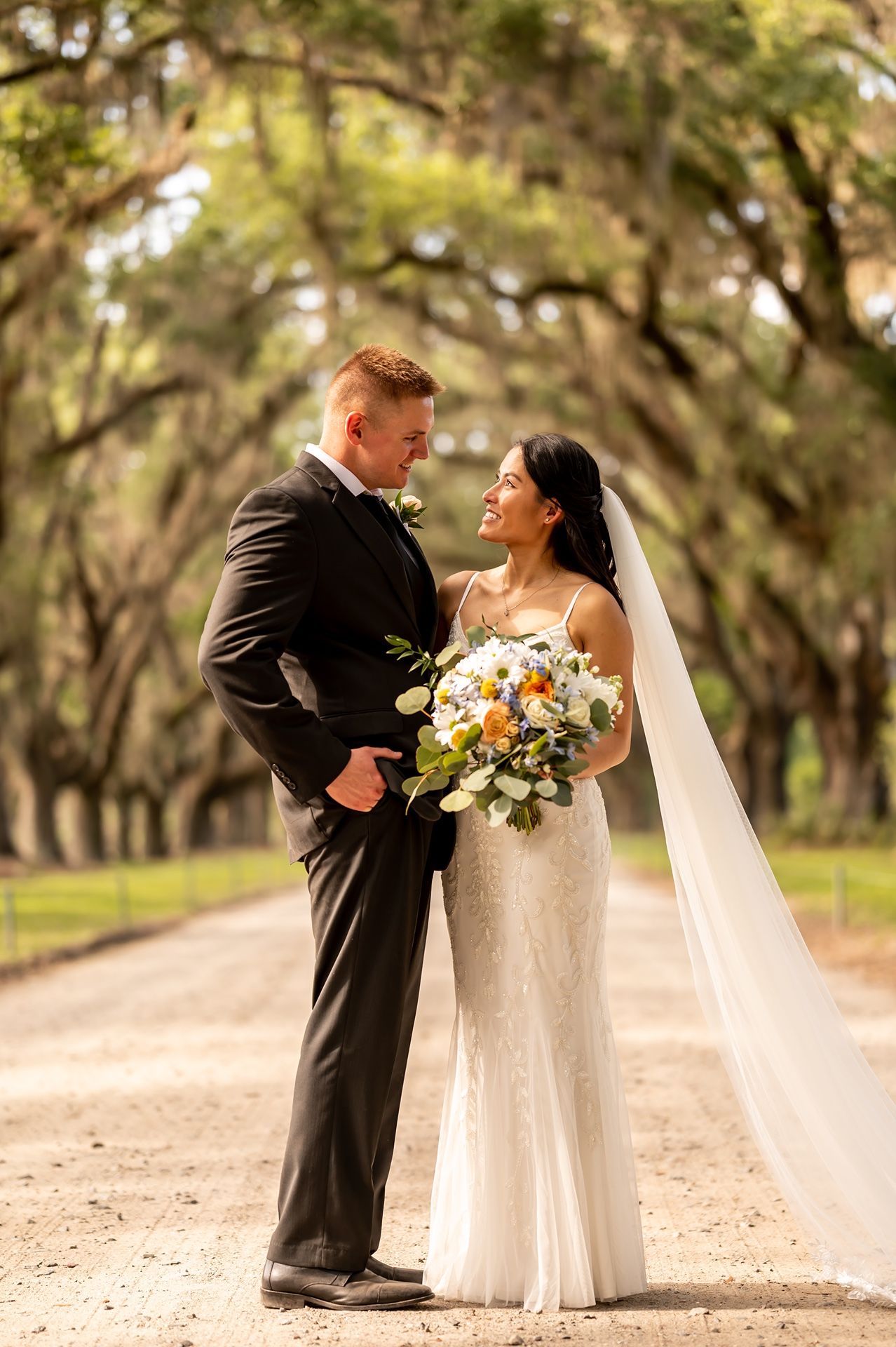 A newlywed couple shares a romantic moment on a tree-lined path with Spanish moss, holding a vibrant floral bouquet.