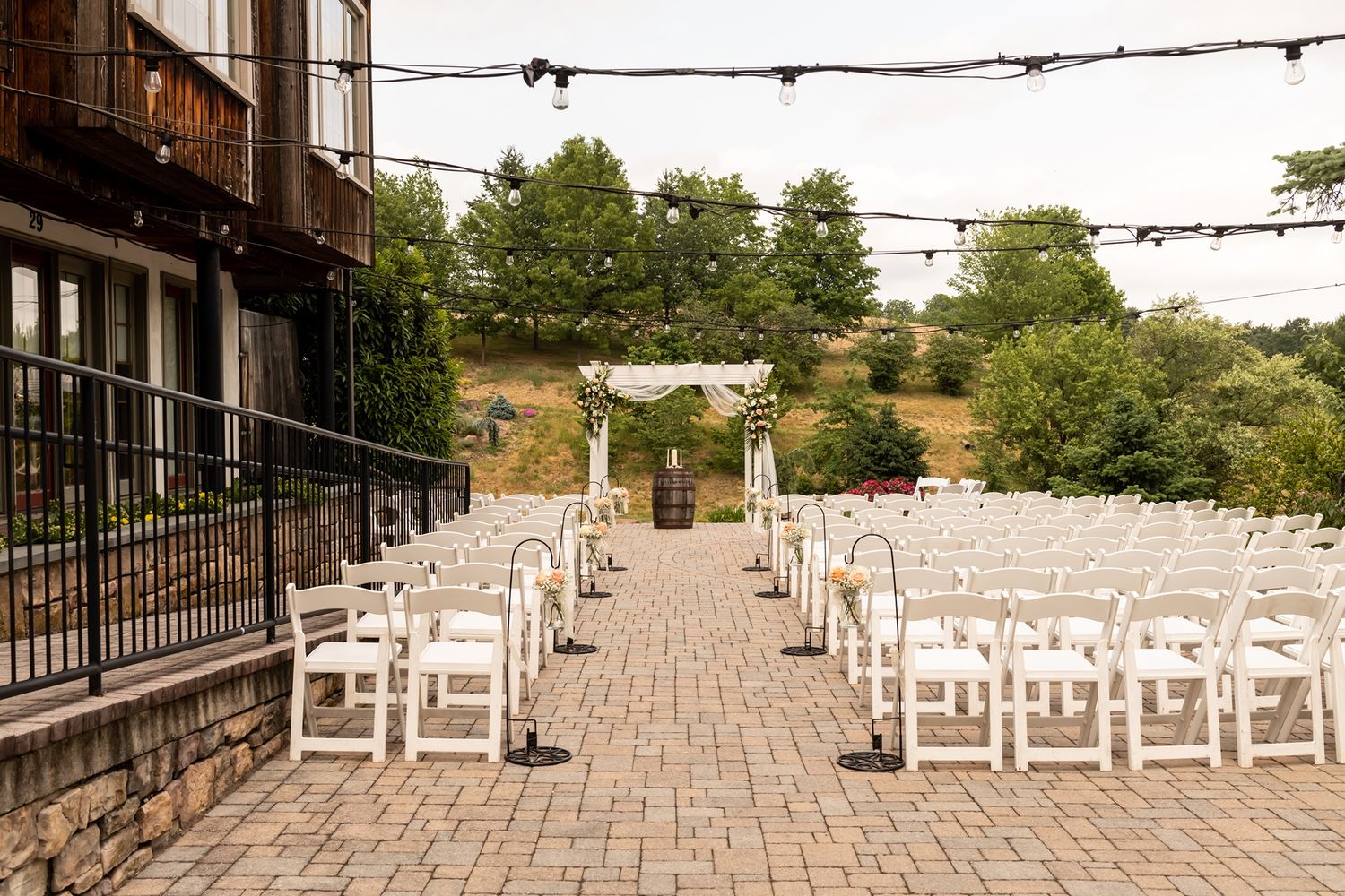 String lights and rows of white folding chairs arranged on a brick patio for an outdoor wedding ceremony.