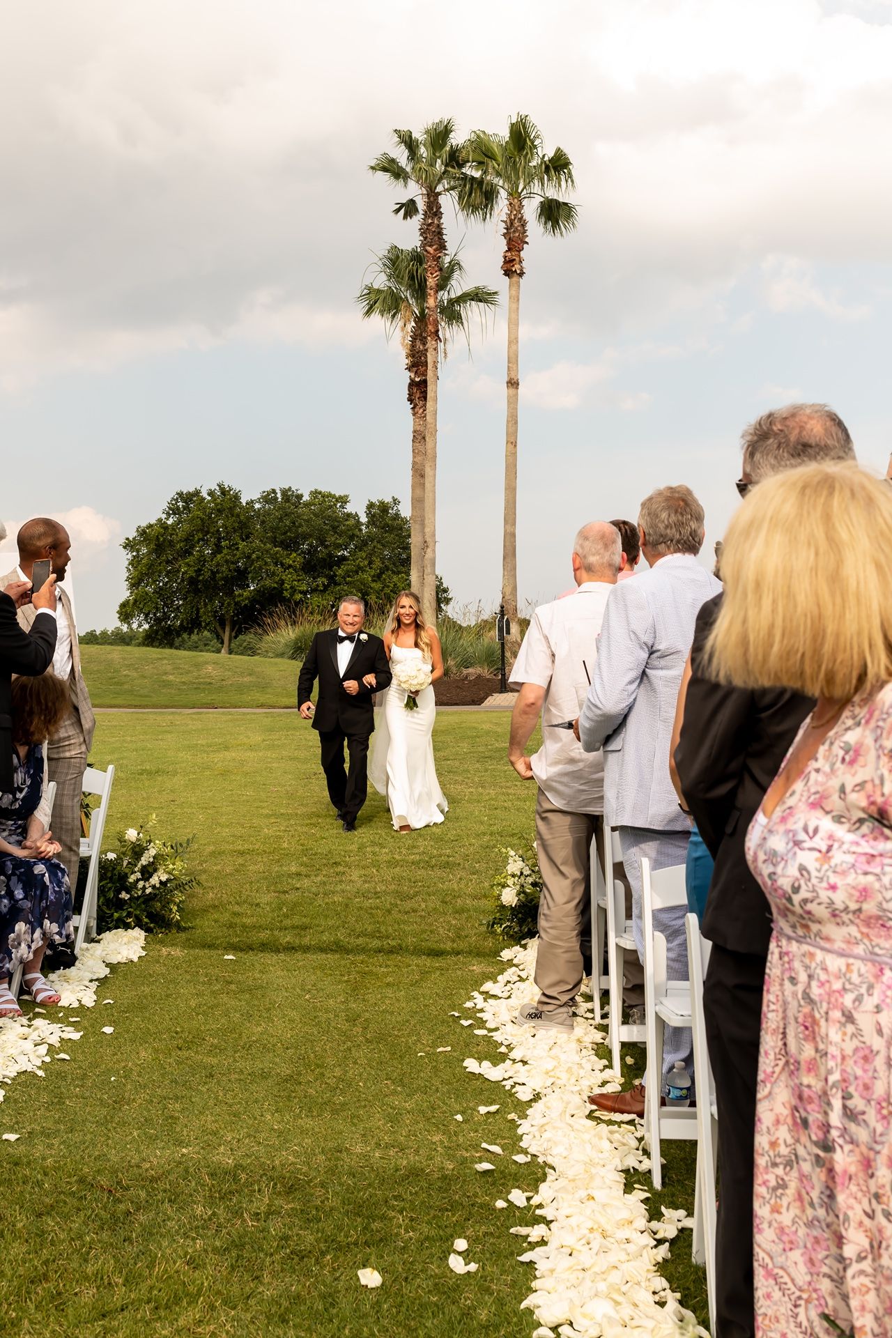 Outdoor wedding ceremony setup with white flower petals lining the aisle and palm trees in the background.