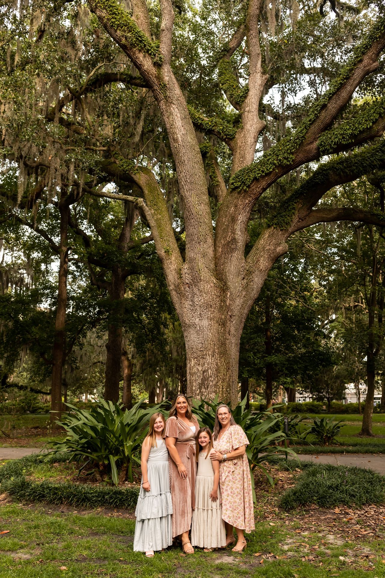 Three individuals in long flowy dresses stand together beneath a majestic old oak tree draped with Spanish moss.