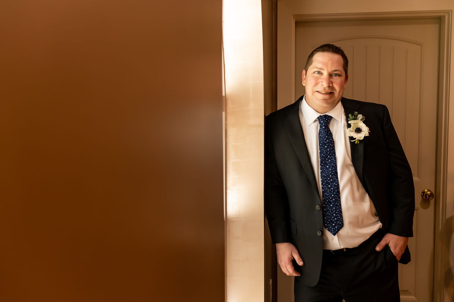 A person wearing a formal black suit and tie stands confidently in a doorway with warm lighting.