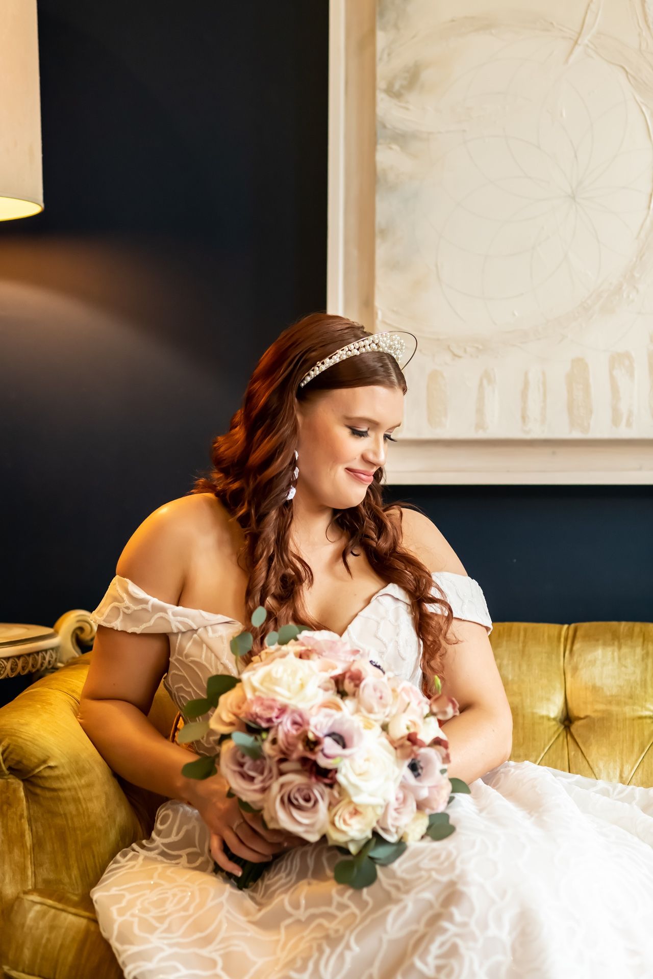 Elegant bride in off-shoulder lace gown sits on yellow velvet sofa holding romantic pink and white floral bouquet.