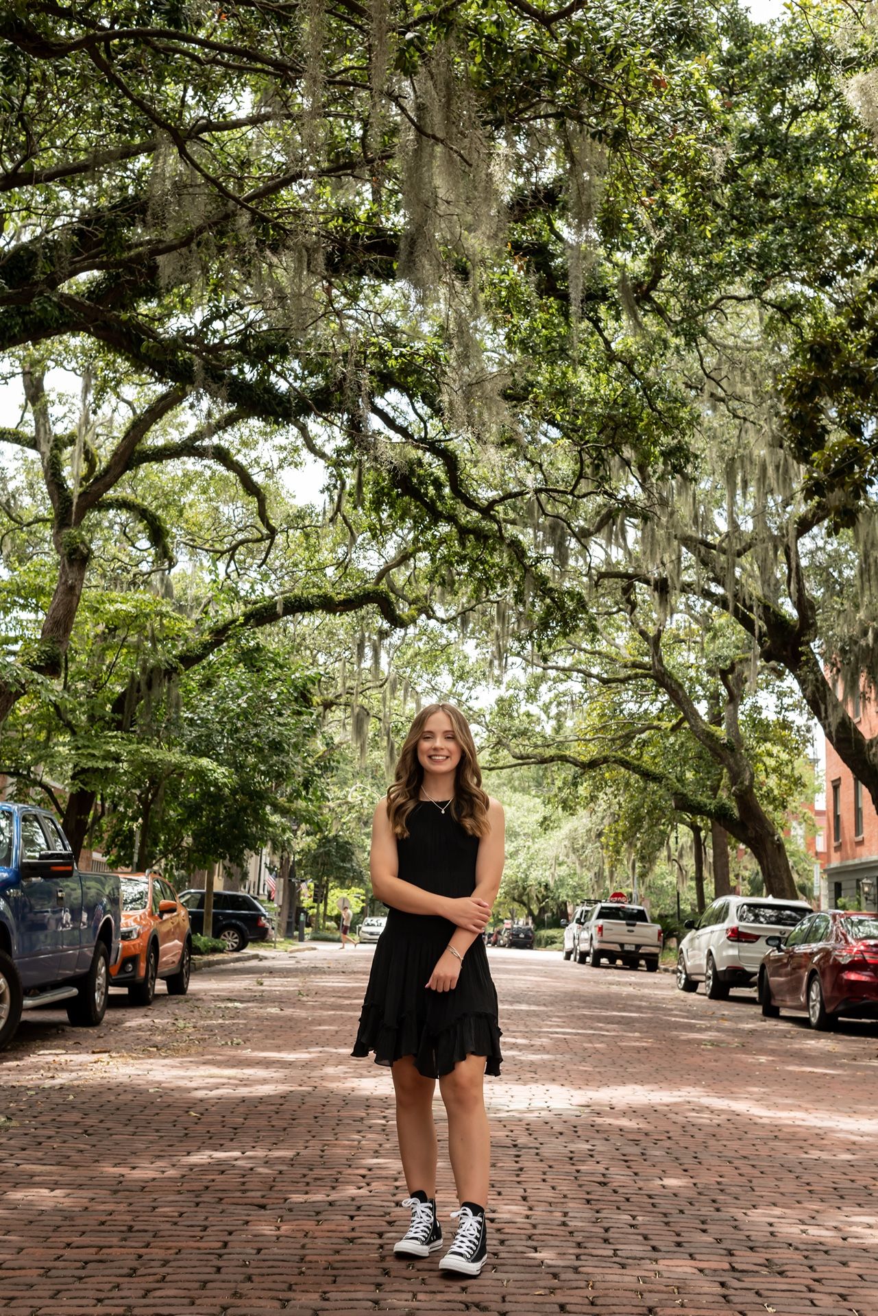 A scenic cobblestone street in Savannah Georgia lined with oak trees draped in Spanish moss and parked cars.
