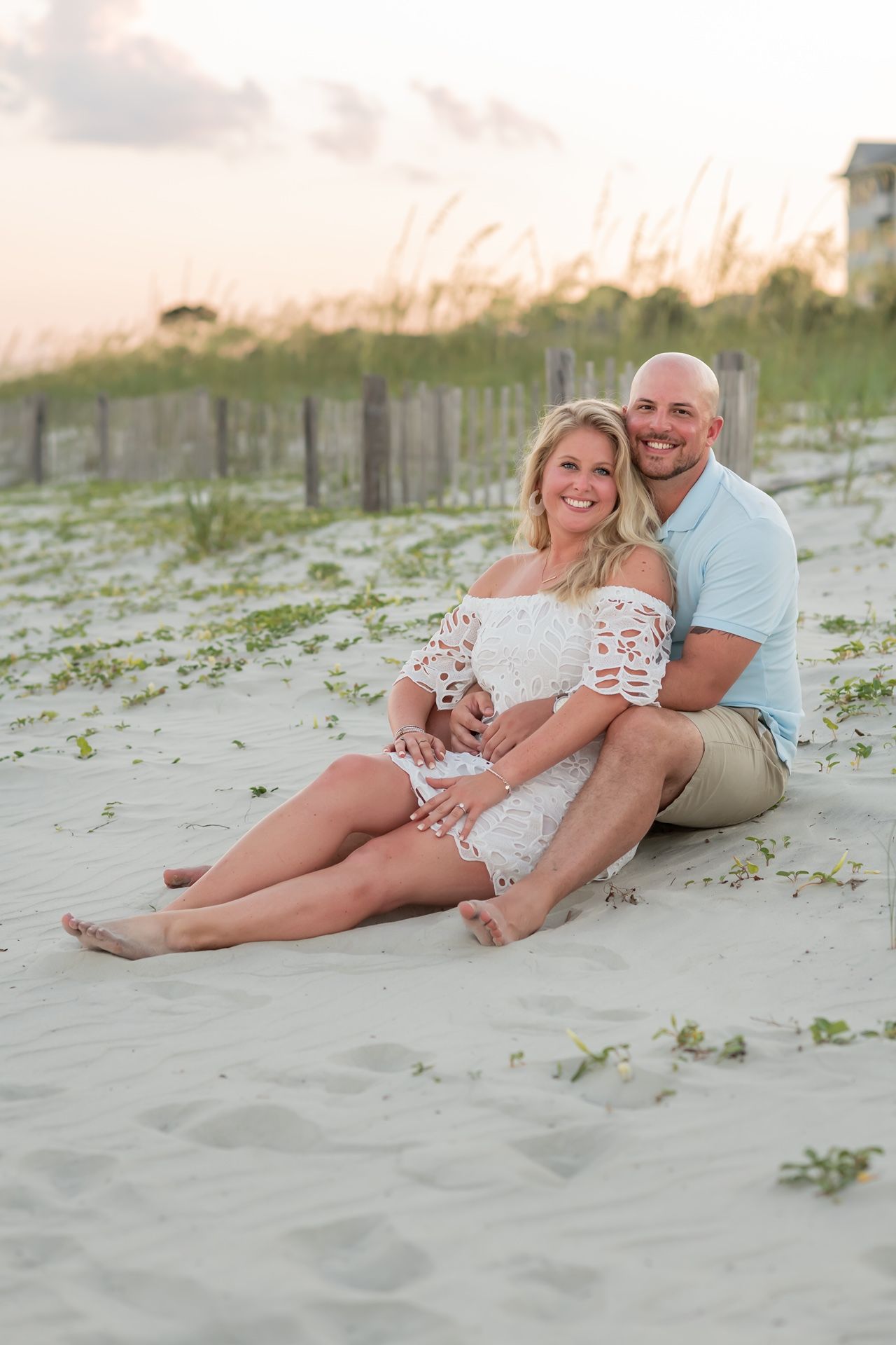 Couple sitting together on sandy beach at sunset with sea oats and dunes in background.