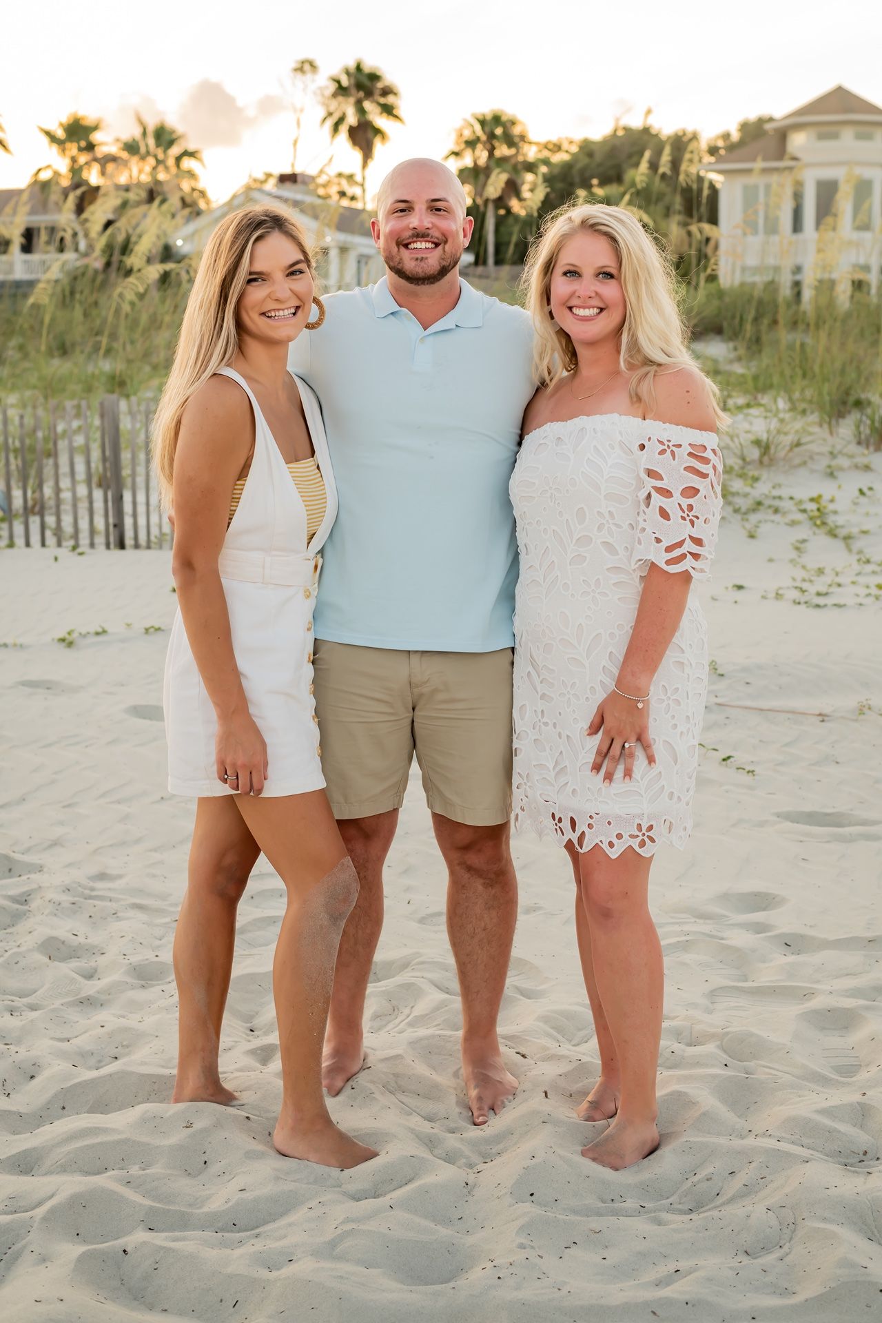 Three people pose together on a sandy beach at sunset with palm trees and a coastal home in the background.