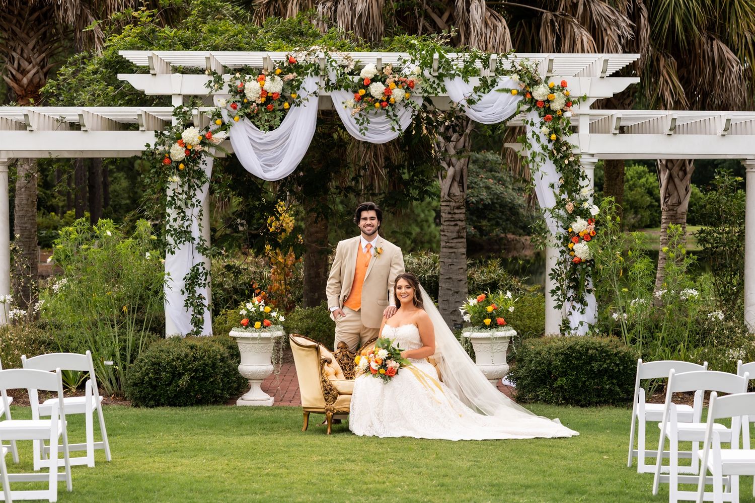 Outdoor garden wedding ceremony setup with white pergola decorated in flowers and fabric draping on manicured lawn.
