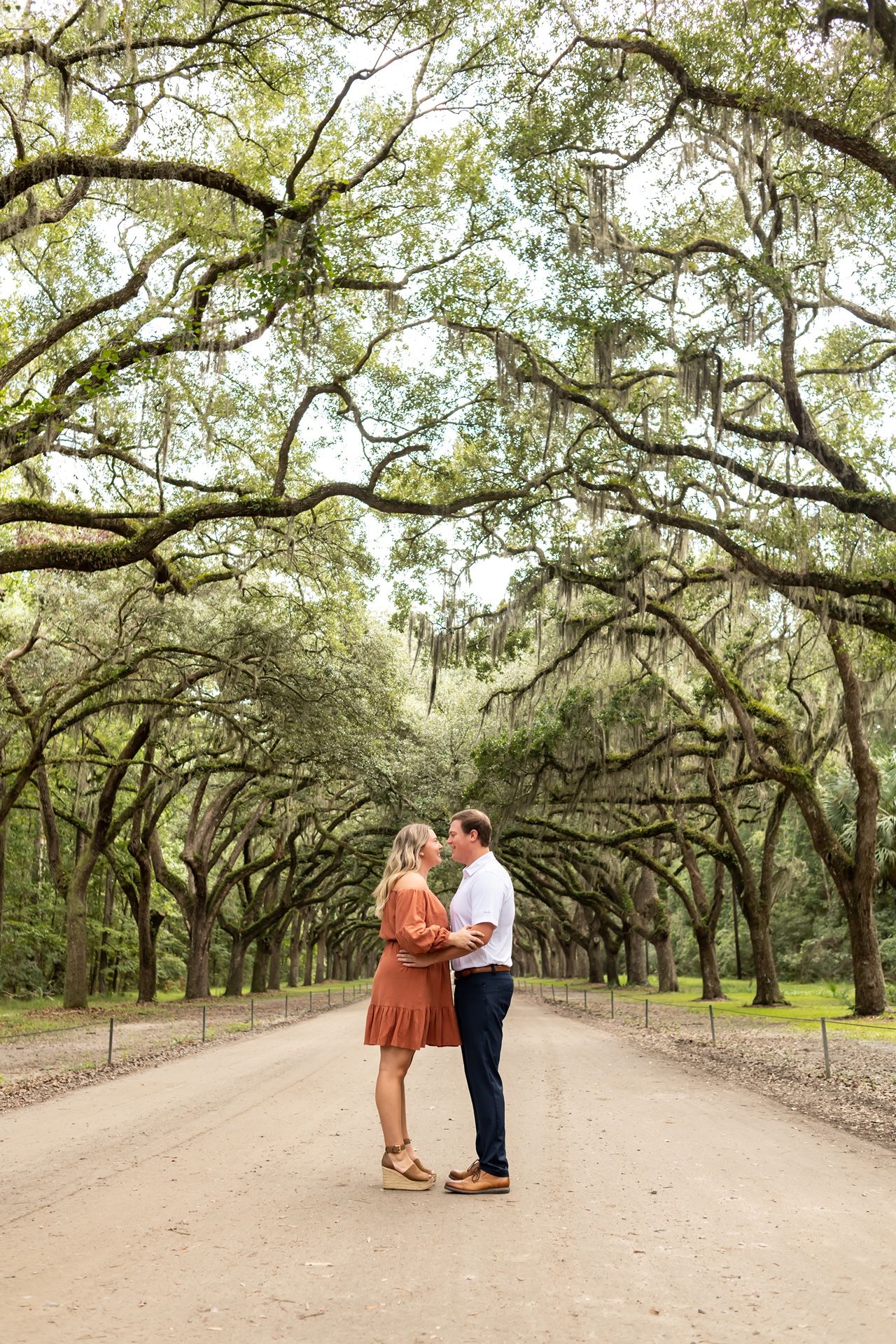 A couple shares a romantic moment beneath Spanish moss-draped oak trees on a dirt path in Savannah, Georgia.