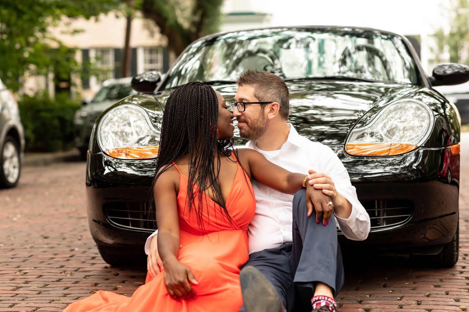 Couple in vintage clothing shares a romantic moment on cobblestone streets while surrounded by classic cars.