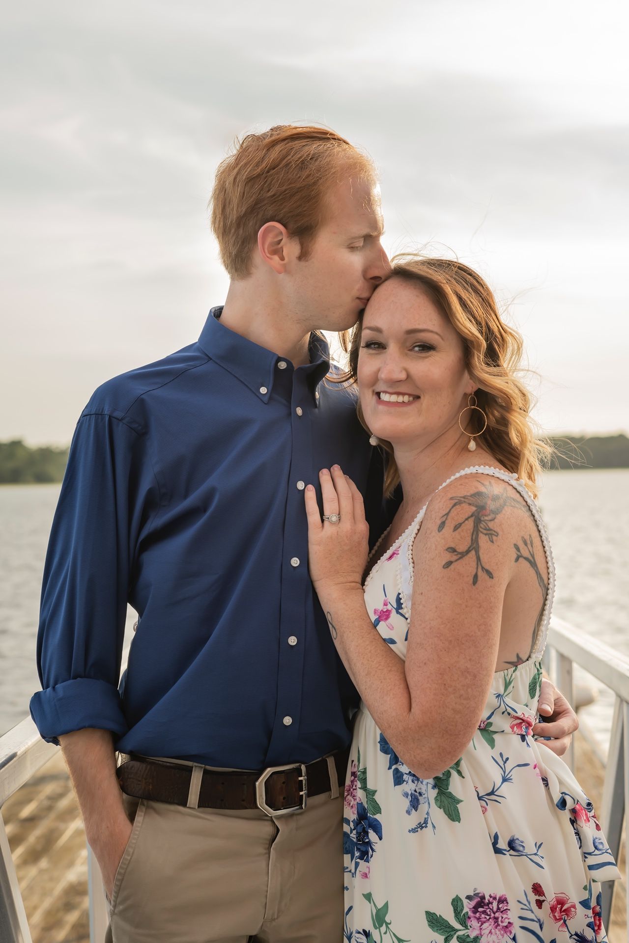 Couple in romantic pose on wooden dock by waterfront with person wearing floral dress and navy shirt.