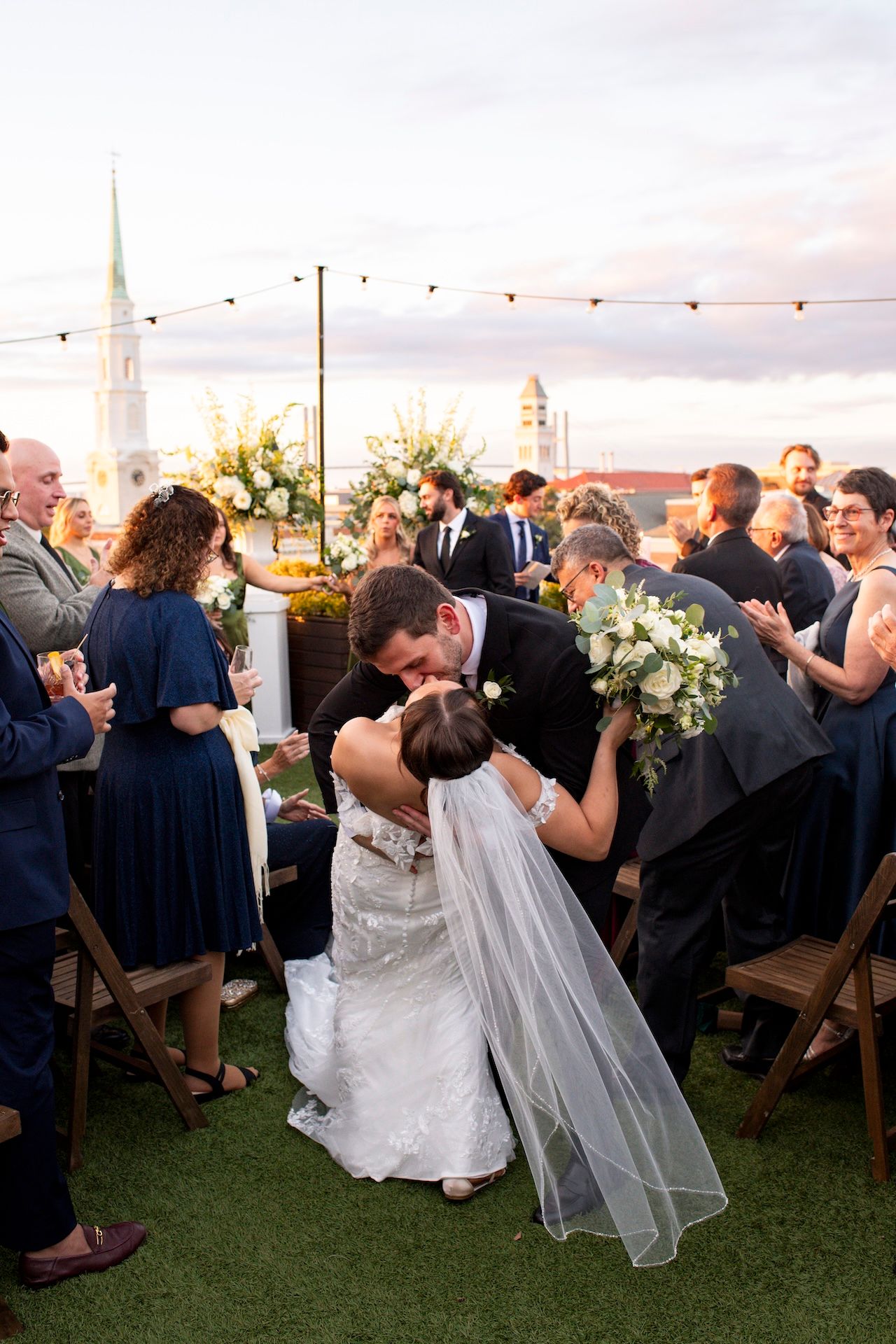 Newlyweds share a romantic dip kiss on an outdoor rooftop terrace during their wedding reception at sunset.