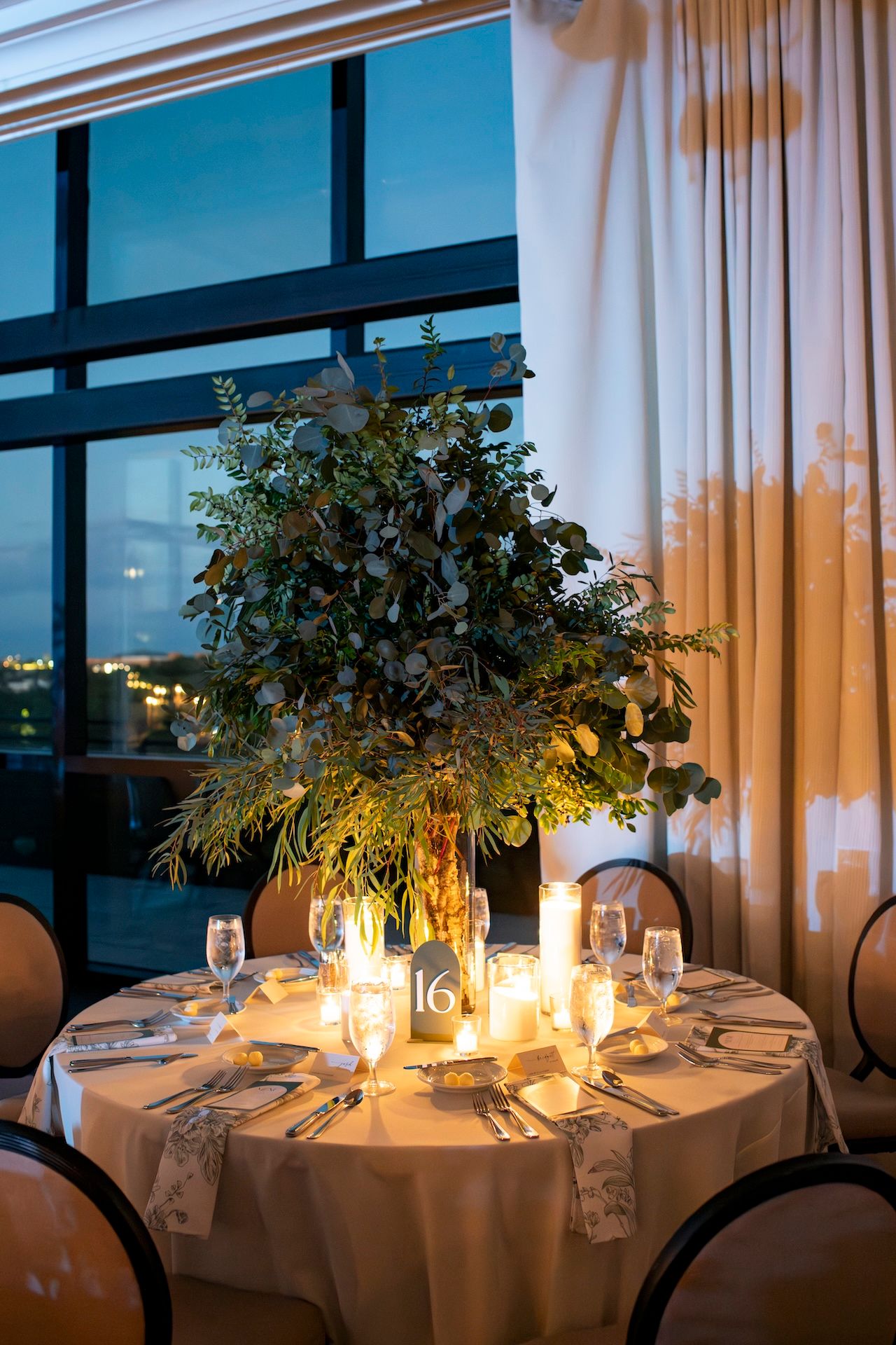 Elegant round dining tables set for dinner with candlelight, white linens, and greenery in a modern restaurant setting.