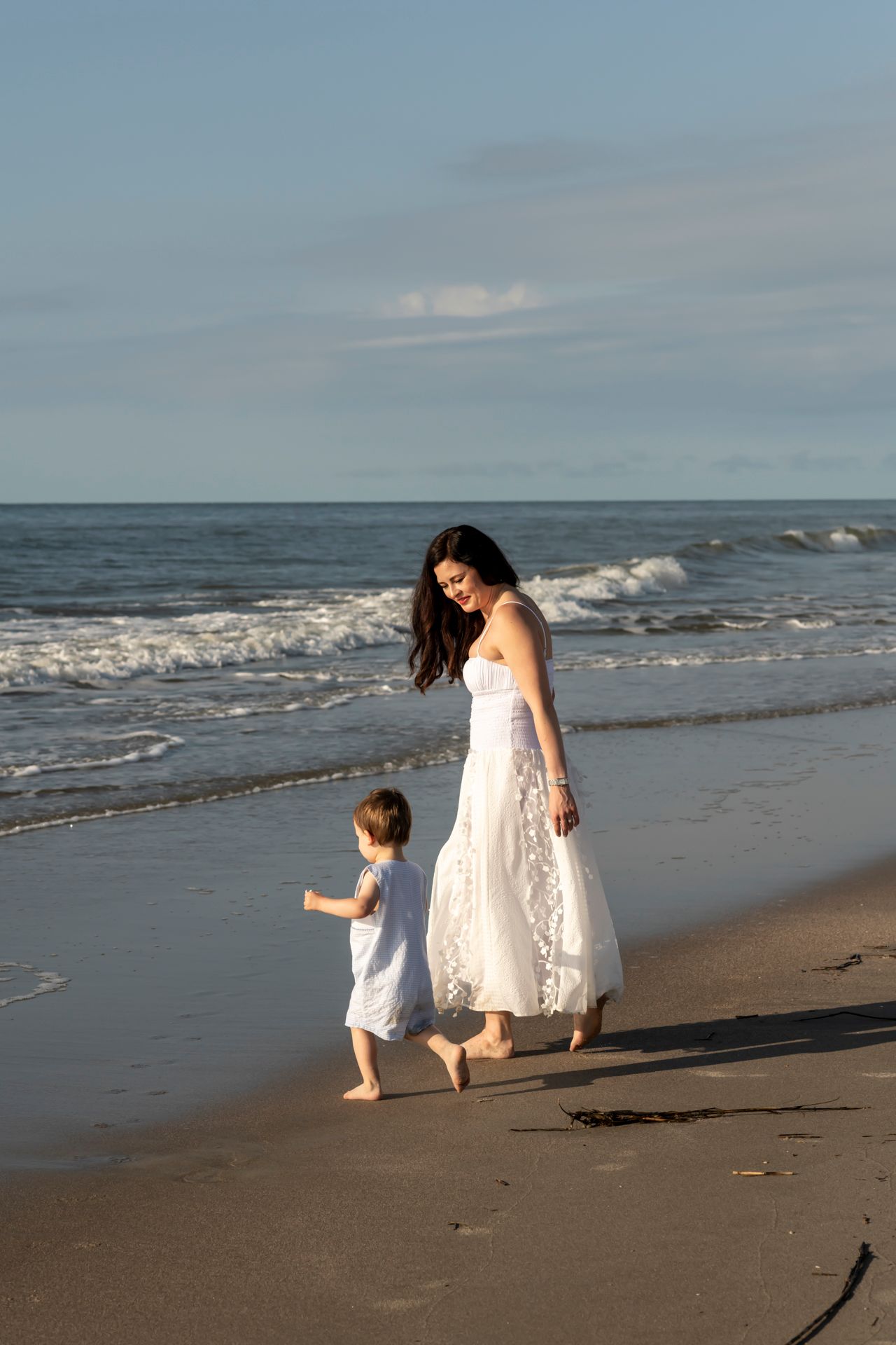 A person in a white dress and child walk together along a scenic beach shoreline at sunset.