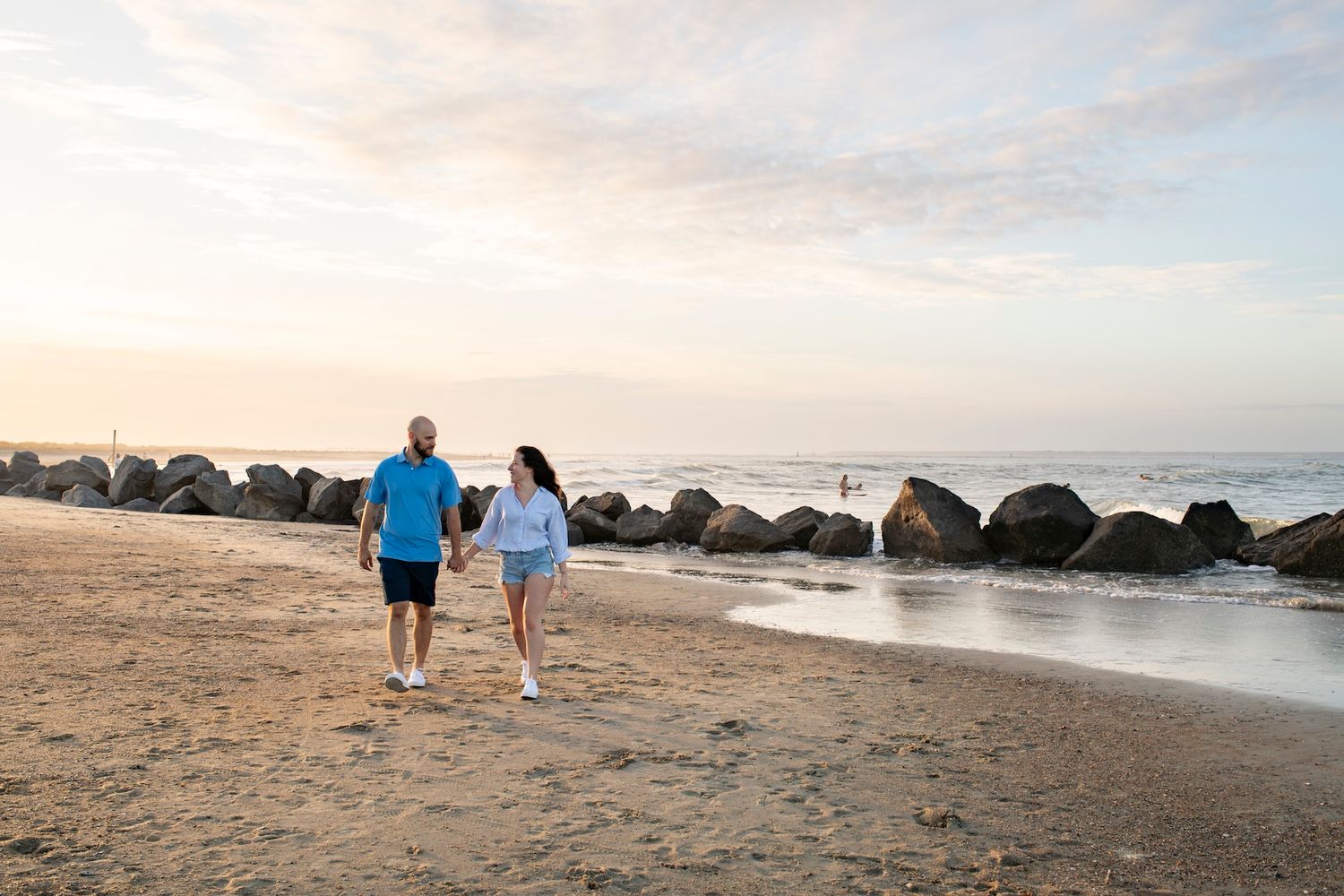 Couple holding hands walking on sandy beach at sunset with large rocks and ocean waves in background.