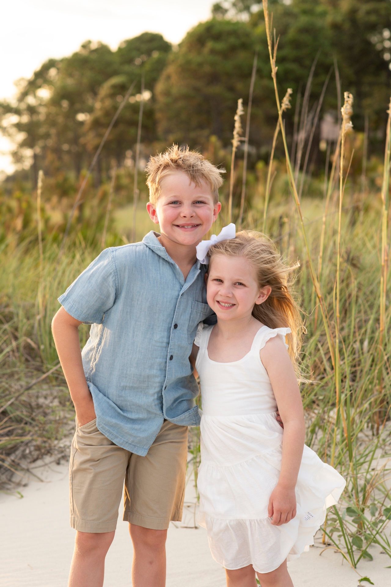 Two smiling children pose on a sandy beach with sea oats, boy in blue shirt and girl in white dress.