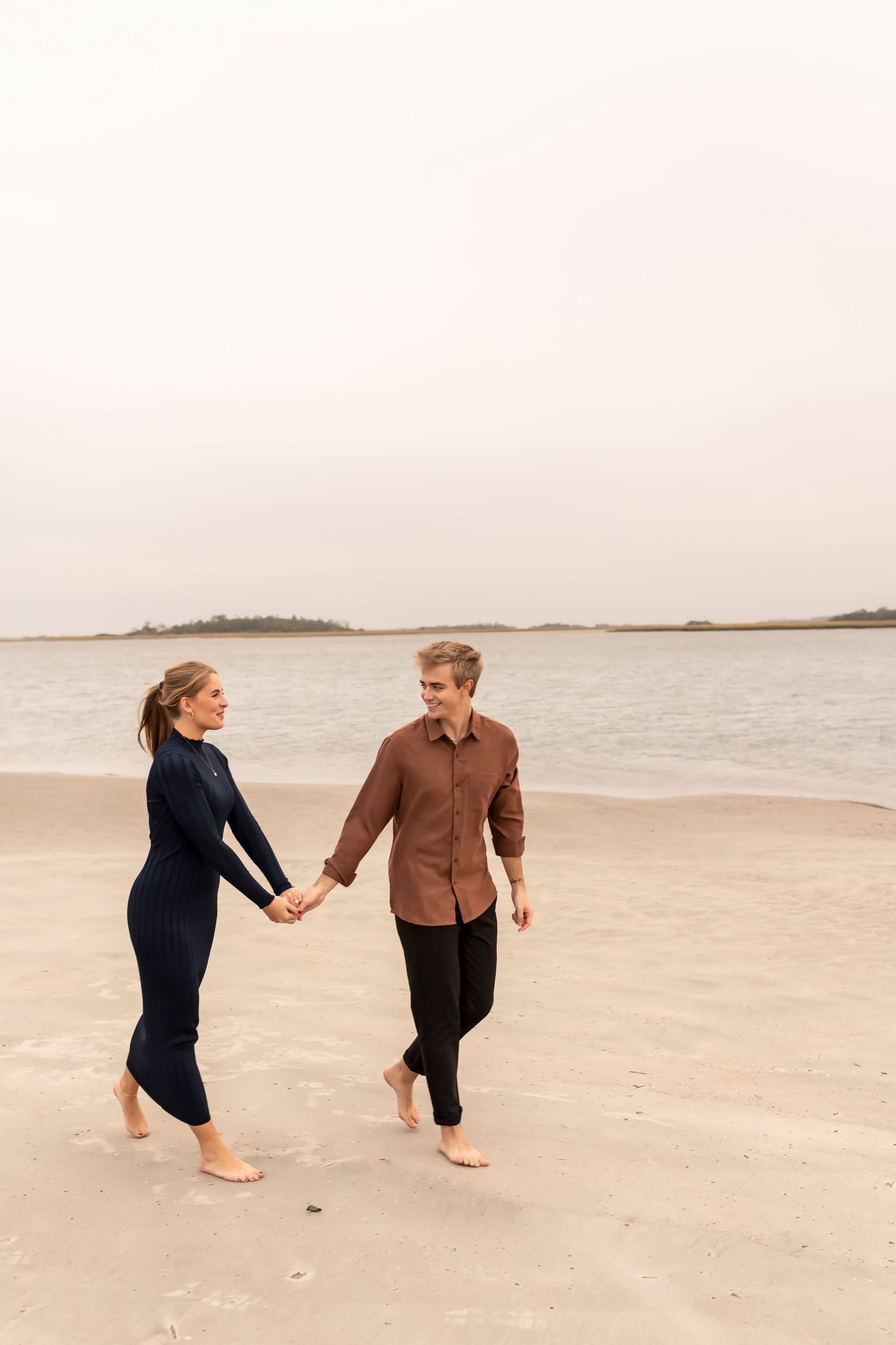 A couple walks hand in hand along a misty beach shoreline at dusk wearing casual attire.