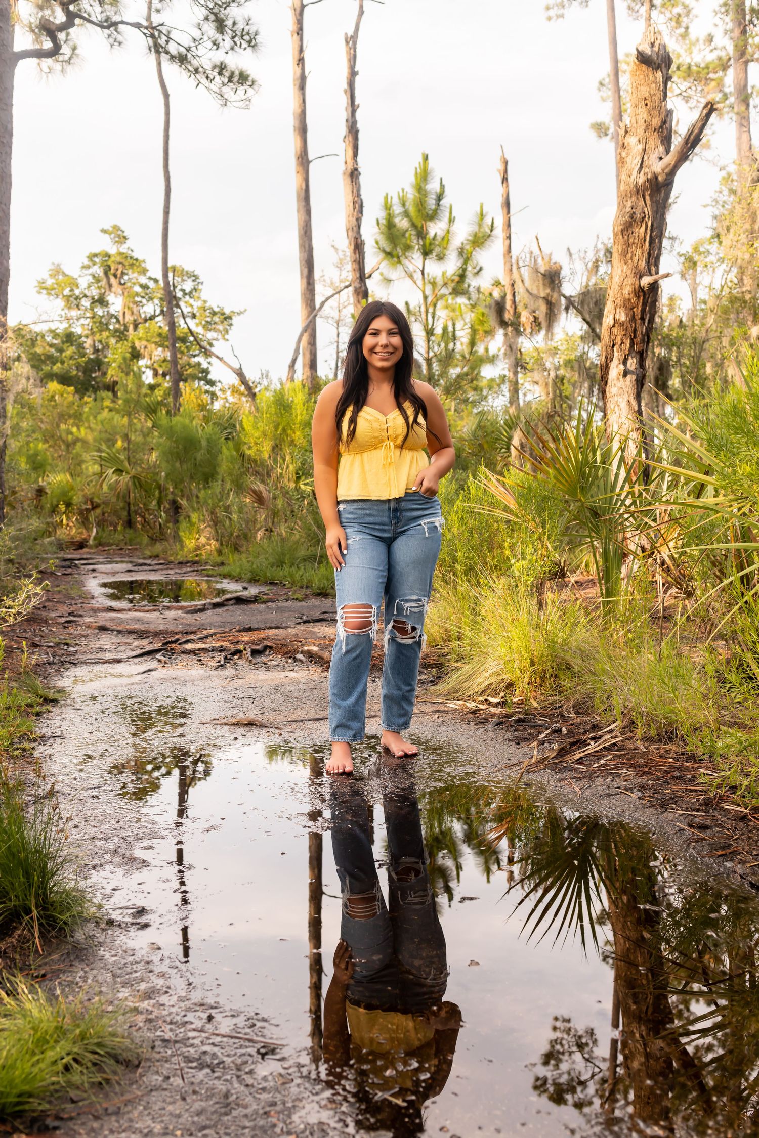 High school senior in yellow top and jeans stands near reflection pool in wooded area.