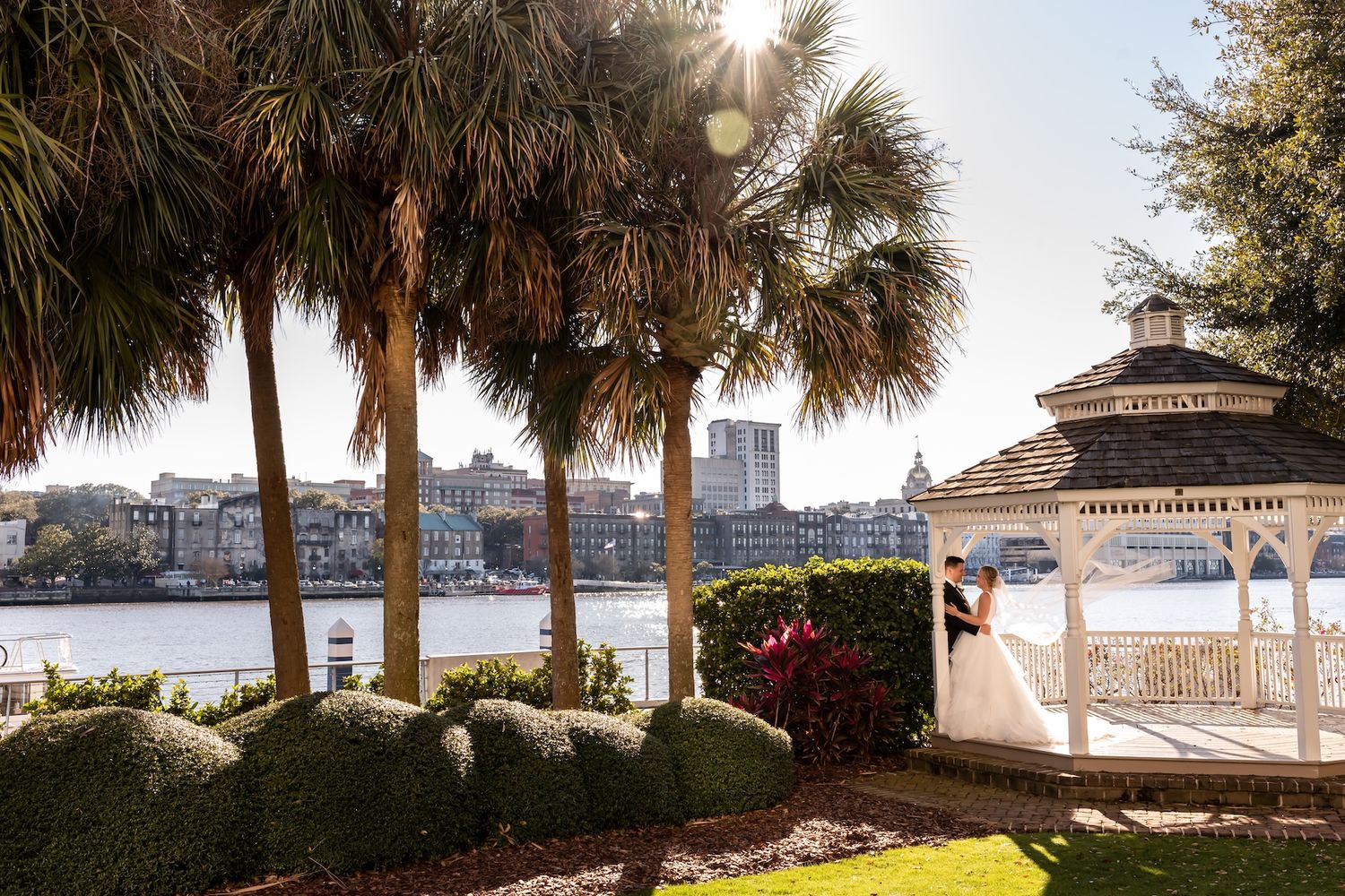 Bride and groom kiss near white gazebo with palm trees and waterfront skyline at sunset.